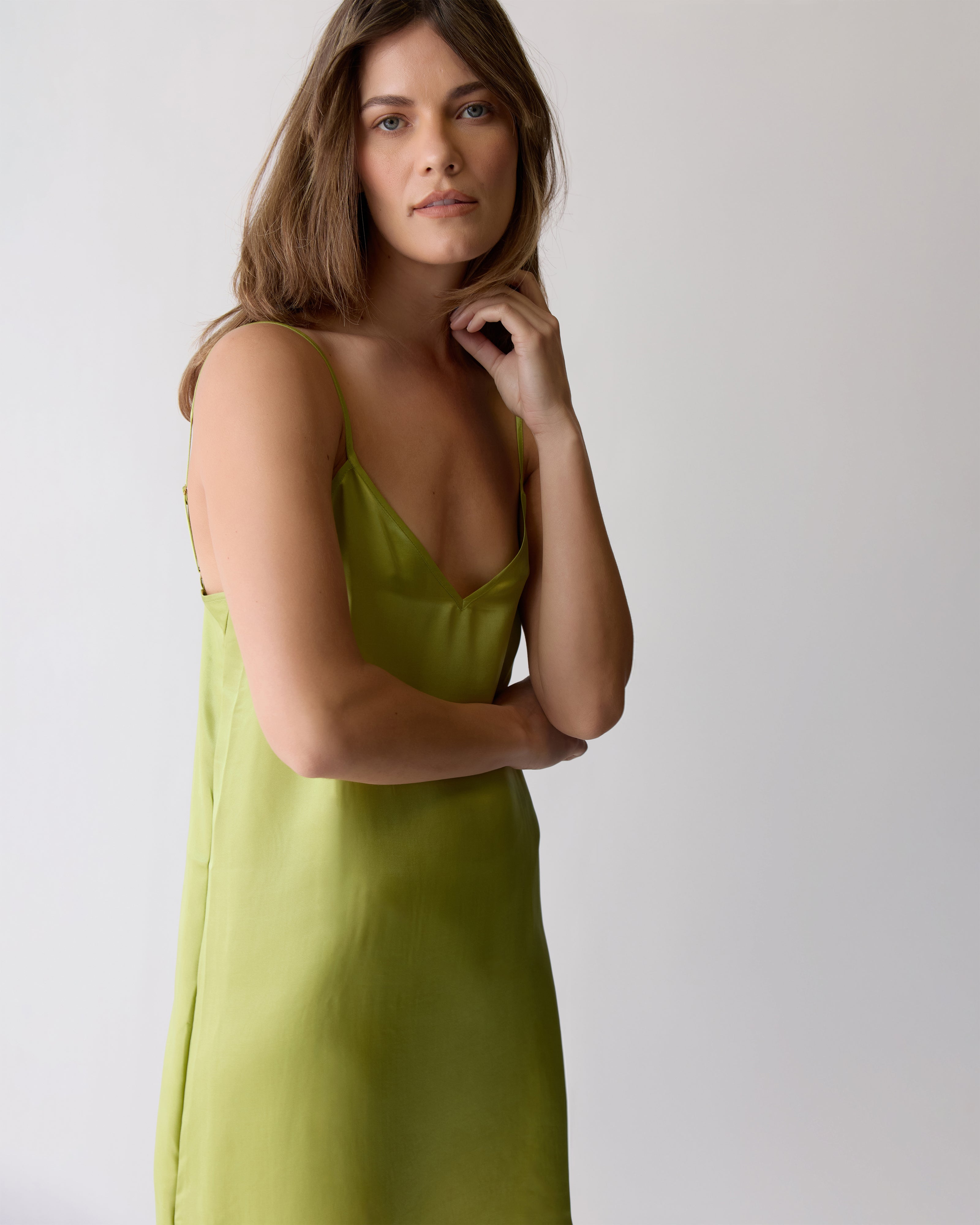 A woman with long brown hair wears The Adonia Satin Slip Dress in lime by Dandy Del Mar, standing against a plain light background.