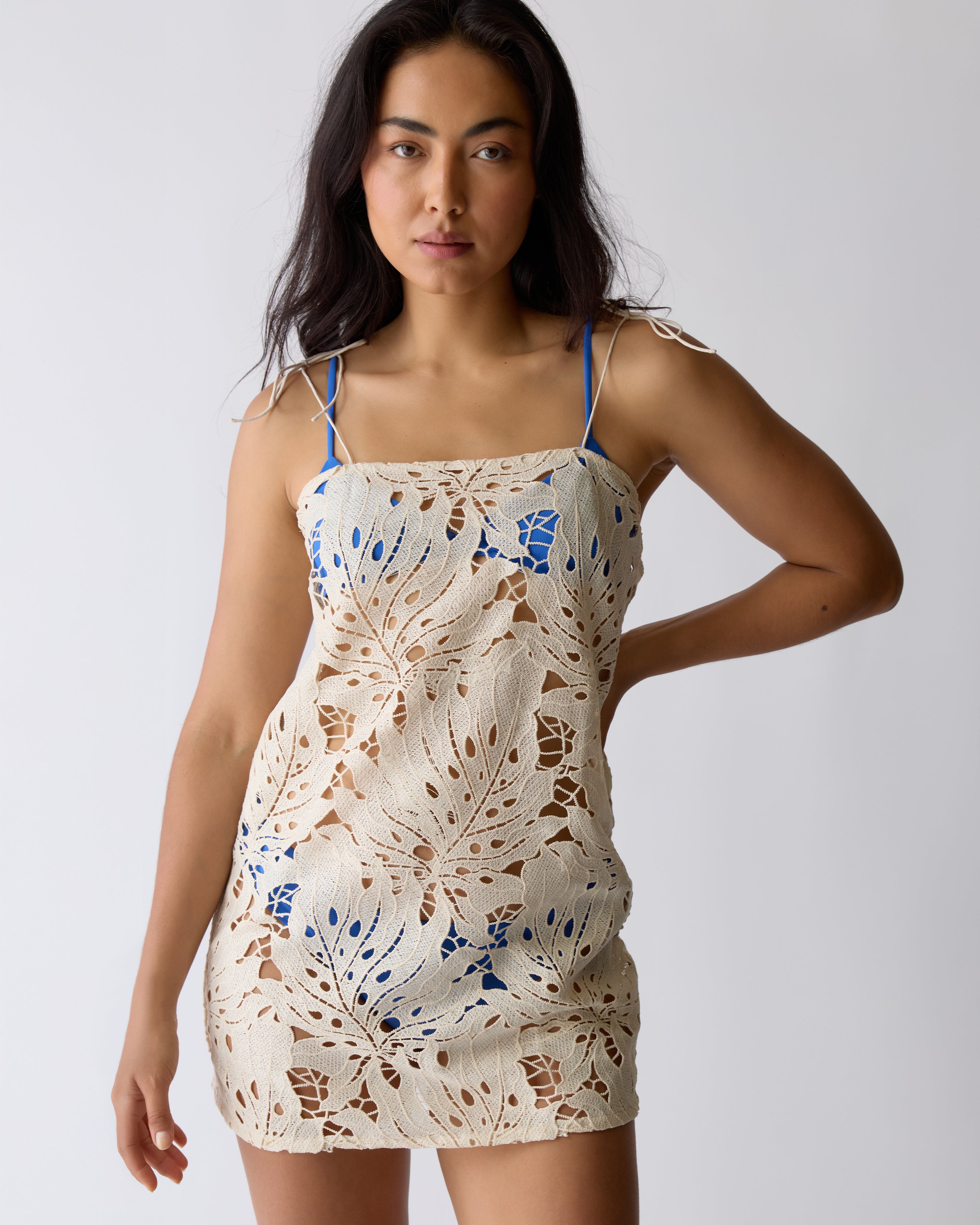 A woman in the Dandy Del Mar Mara Tie Strap Dress - Scallop stands against a plain light background, featuring beige scalloped crochet over blue undergarments.