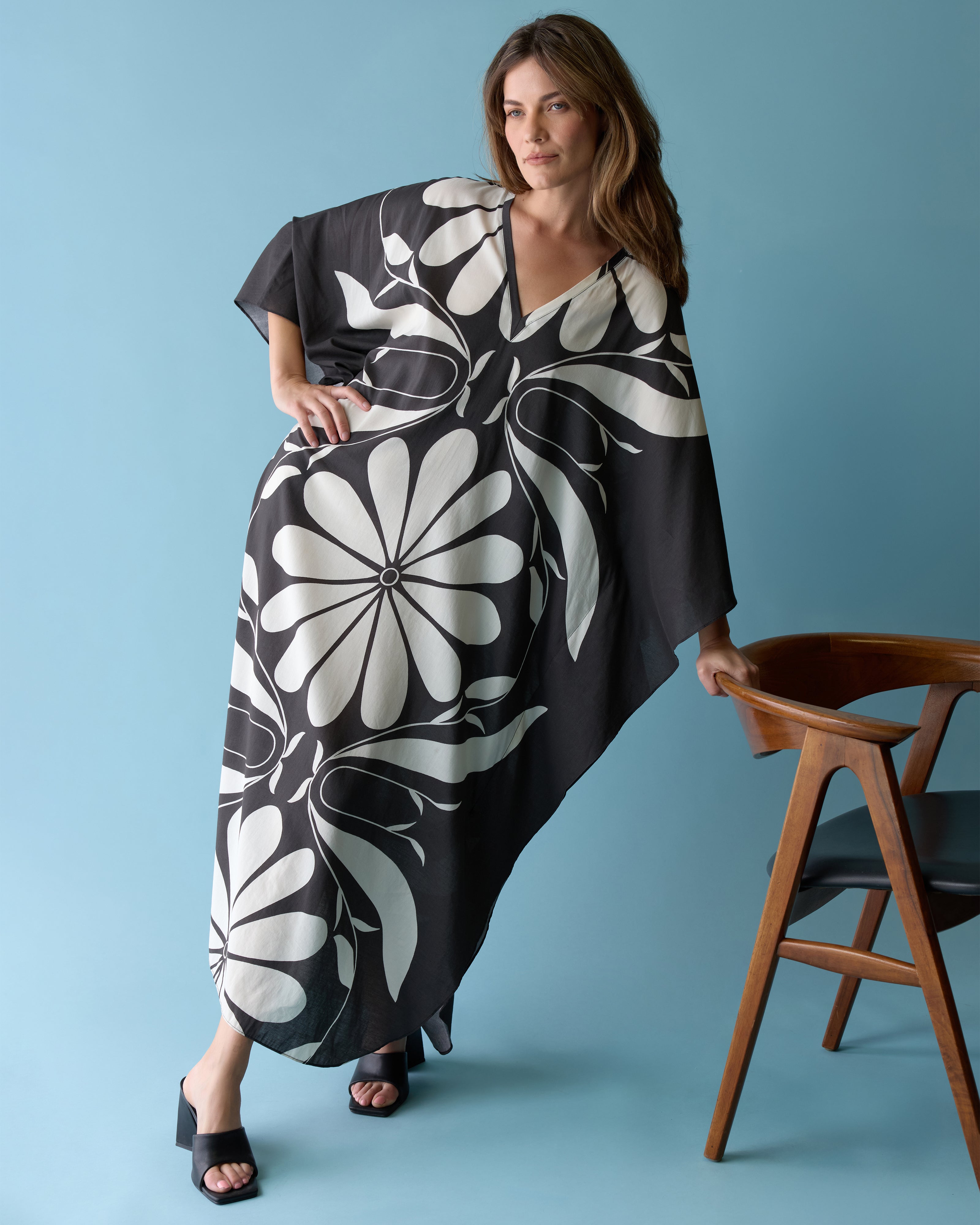 A woman in The Tropea Kaftan - Albatross by Dandy Del Mar stands against a light blue background, leaning on a wooden chair.