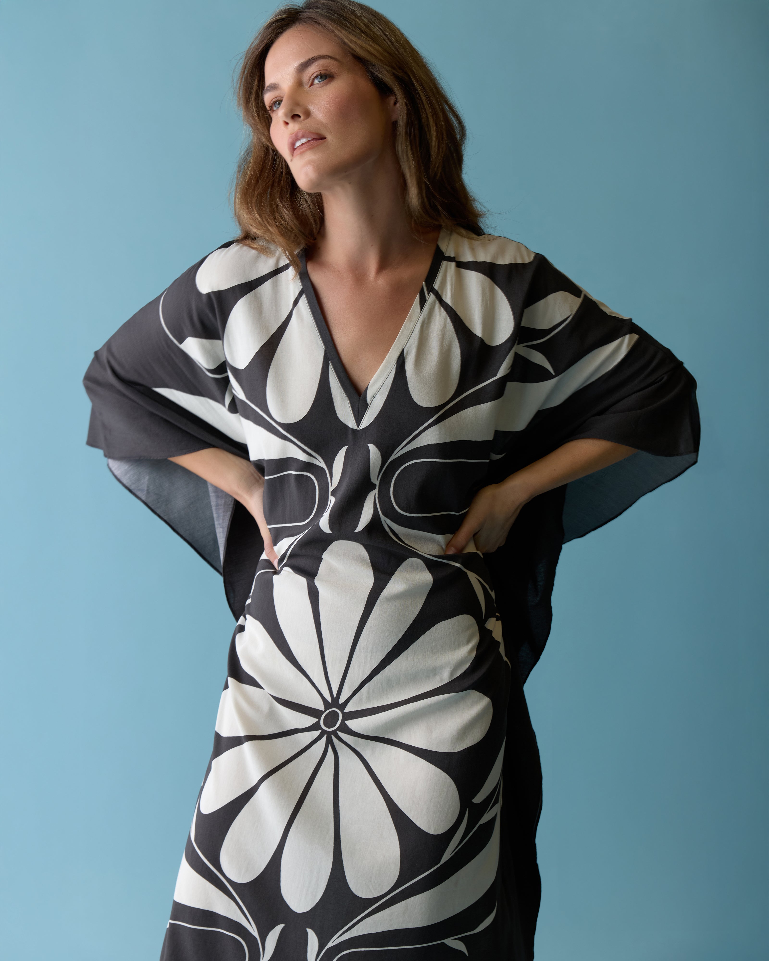 A woman models The Tropea Kaftan - Albatross by Dandy Del Mar, featuring a bold black and white floral print, while standing against a plain light blue background and looking slightly upward.