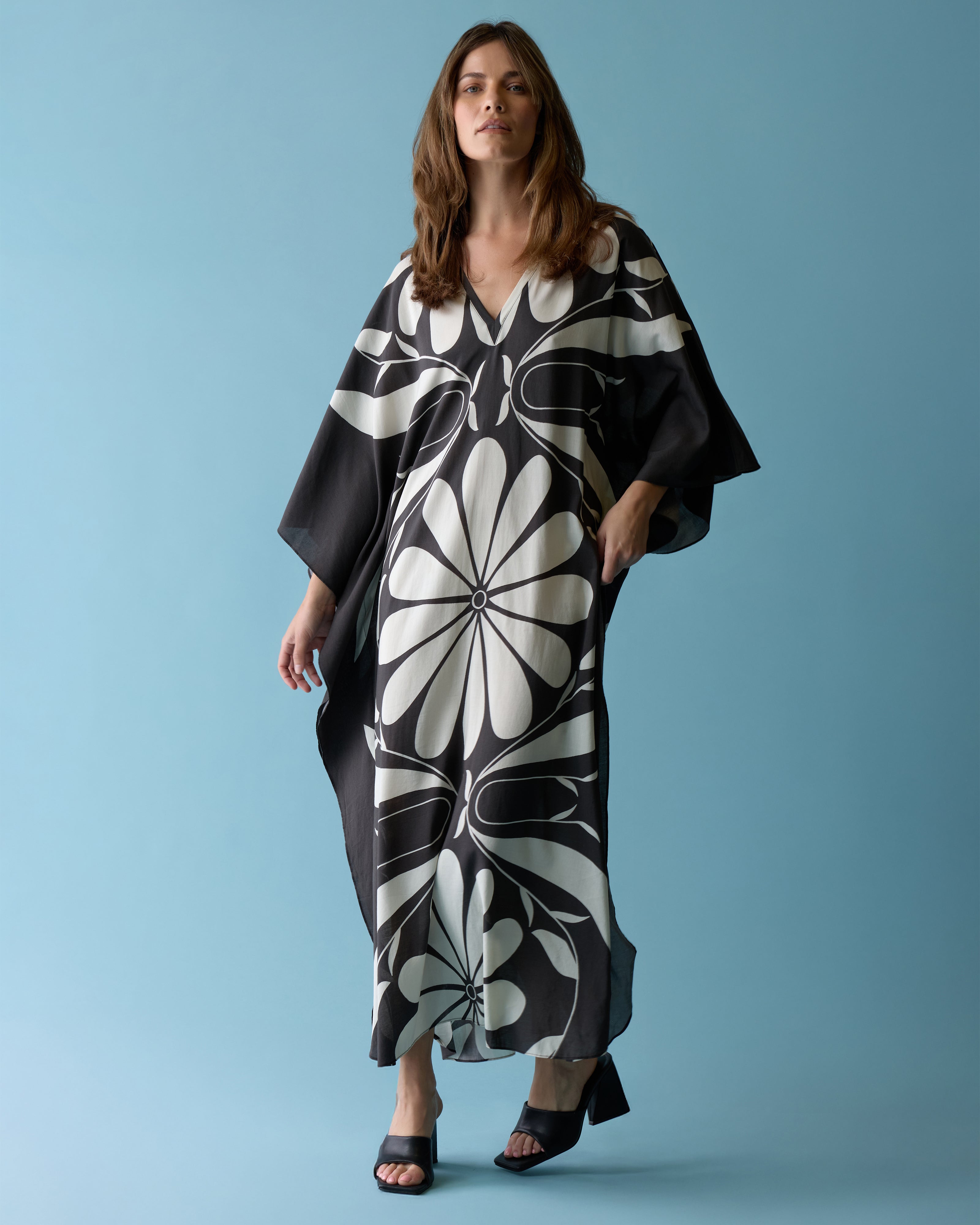 A woman stands against a blue background wearing Dandy Del Mar's The Tropea Kaftan - Albatross and black heeled sandals.