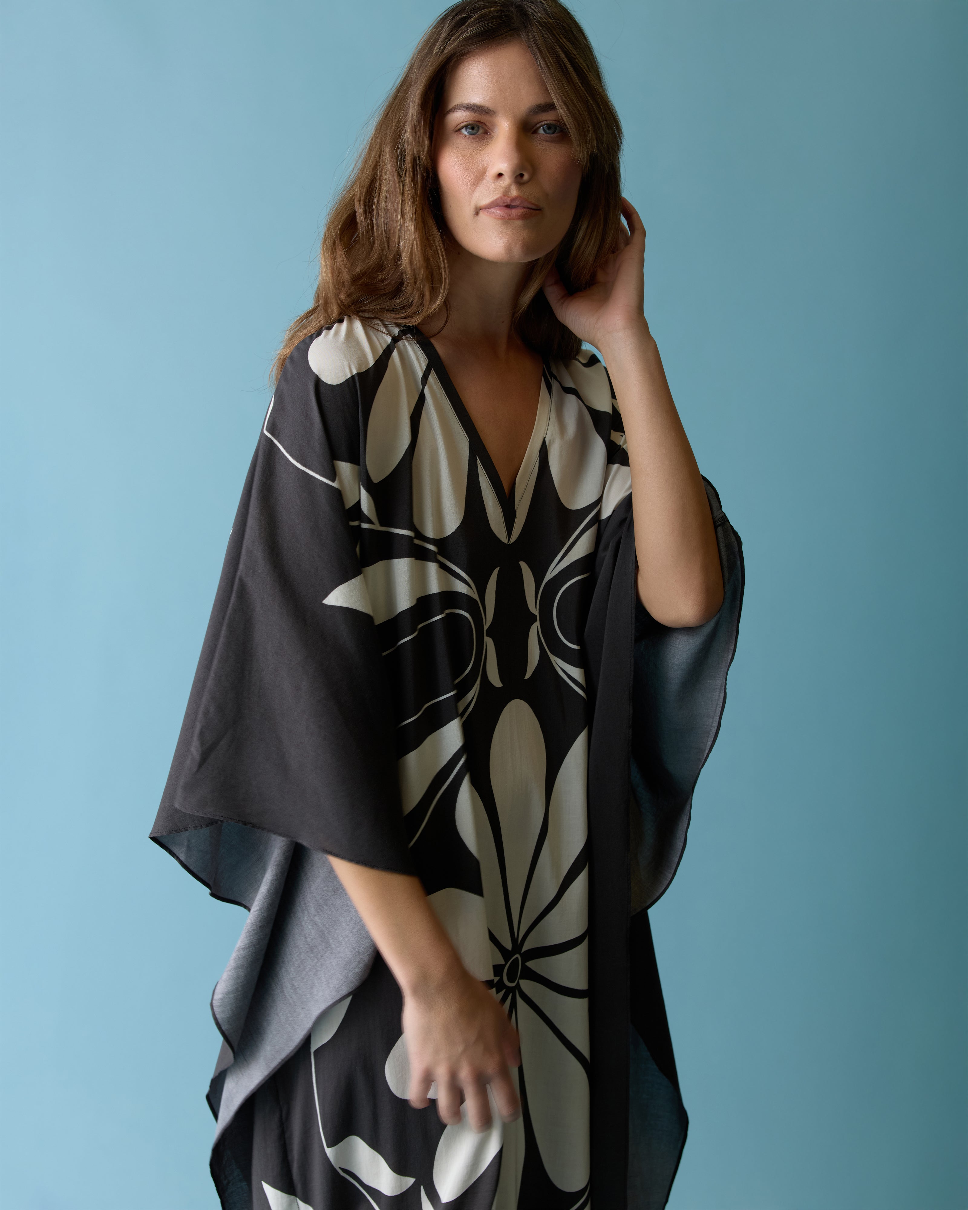 A woman stands against a blue background wearing The Tropea Kaftan - Albatross by Dandy Del Mar, featuring a vivid print and wide sleeves, gazing at the camera.