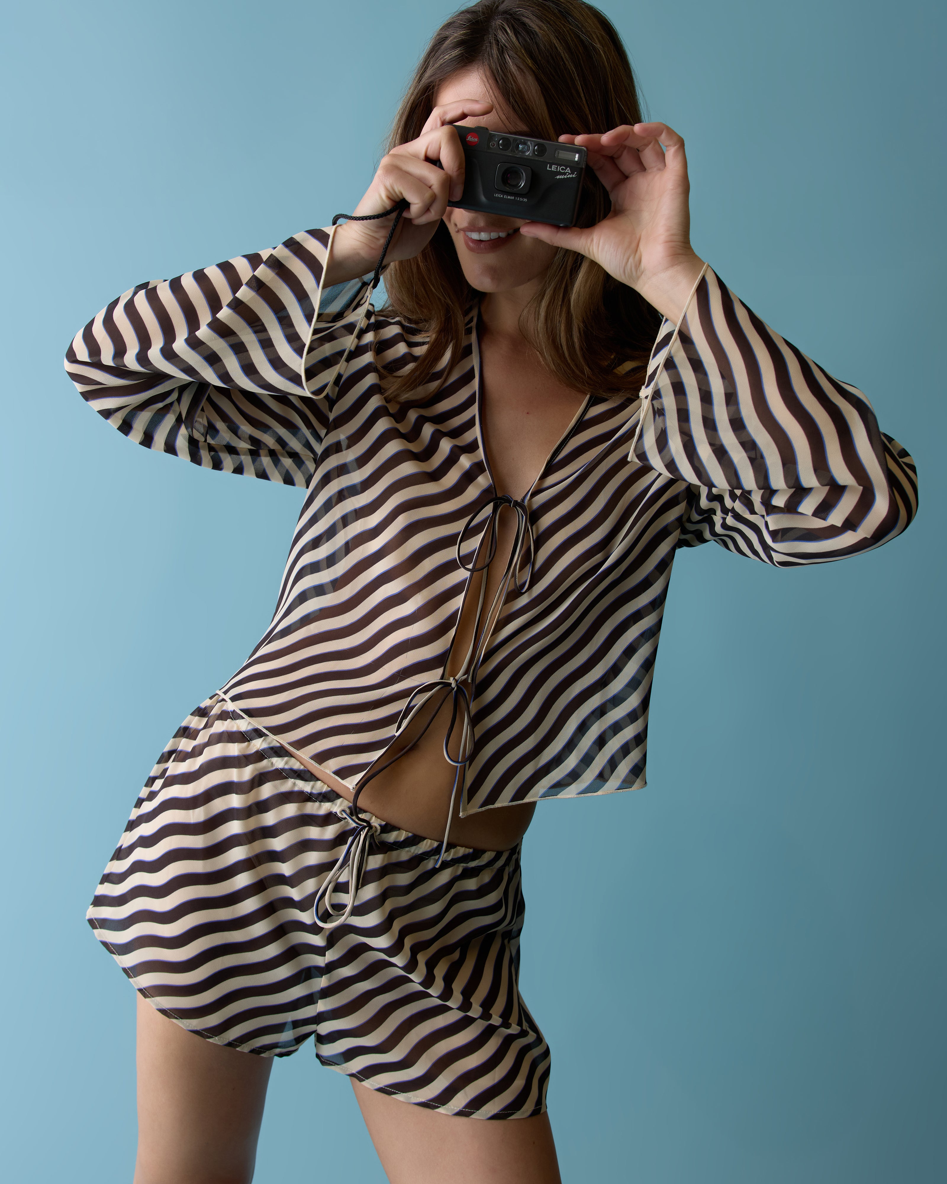 Wearing The Curacao Short - Tierra by Dandy Del Mar, a person poses against a blue background in wavy striped relaxed fit top and chiffon shorts with an elastic waistband, holding a camera up for a photo.