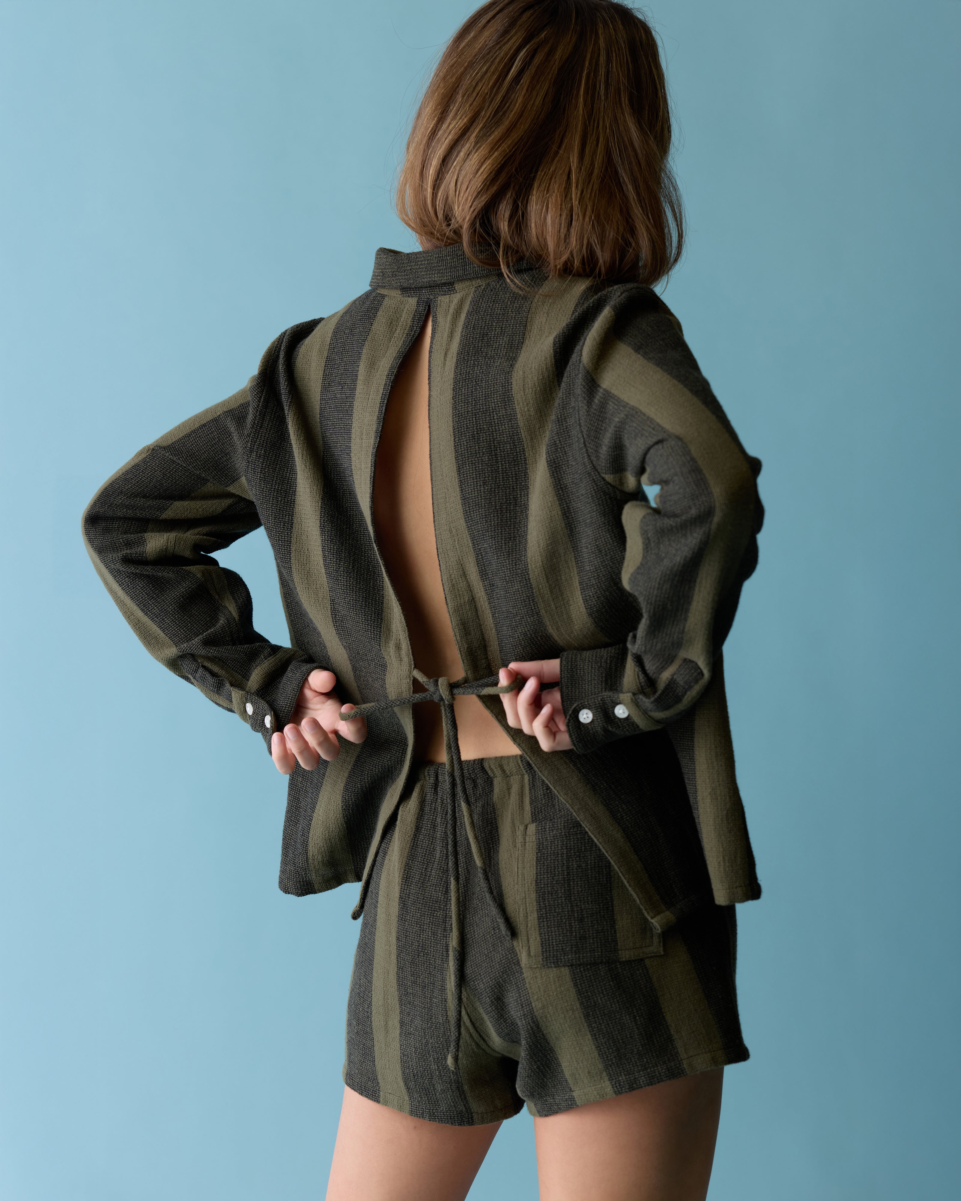 A person stands against a blue background wearing Dandy Del Mar’s The Sevilla Top – Onyx, a long-sleeve, backless shirt tied at the waist with stripes and matching shorts made from yarn-dyed woven fabric.
