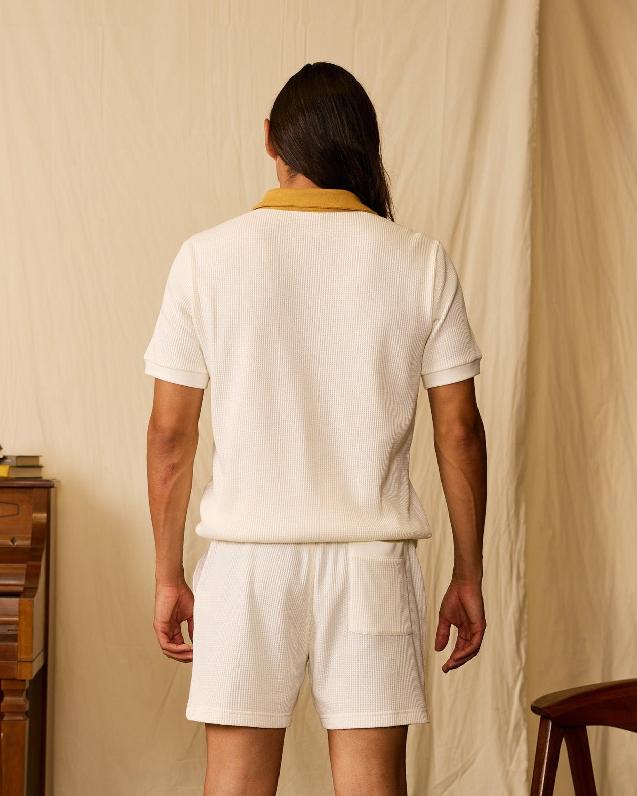 A person with long hair stands facing away, wearing Dandy Del Mar's The Cannes Waffle Knit Polo Shirt in Vintage Ivory, paired with matching white shorts, set against a beige background.
