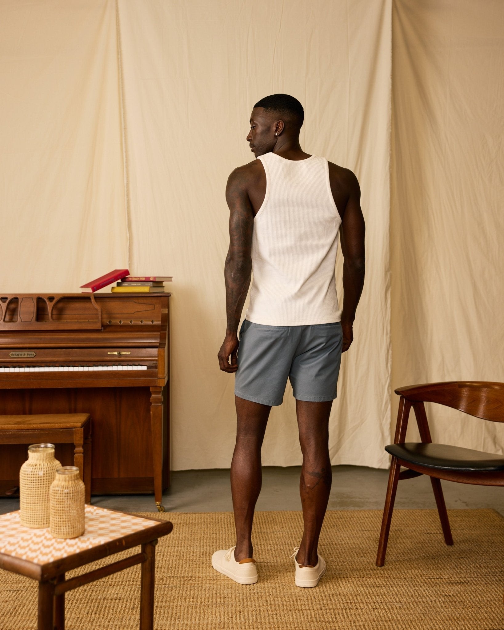 A man faces away, wearing a white tank top and Dandy Del Mar’s The Rhodes Twill Shorts in Abyss. He’s in a room with a piano, stacked books, a wooden chair, and a small table with wicker decor.