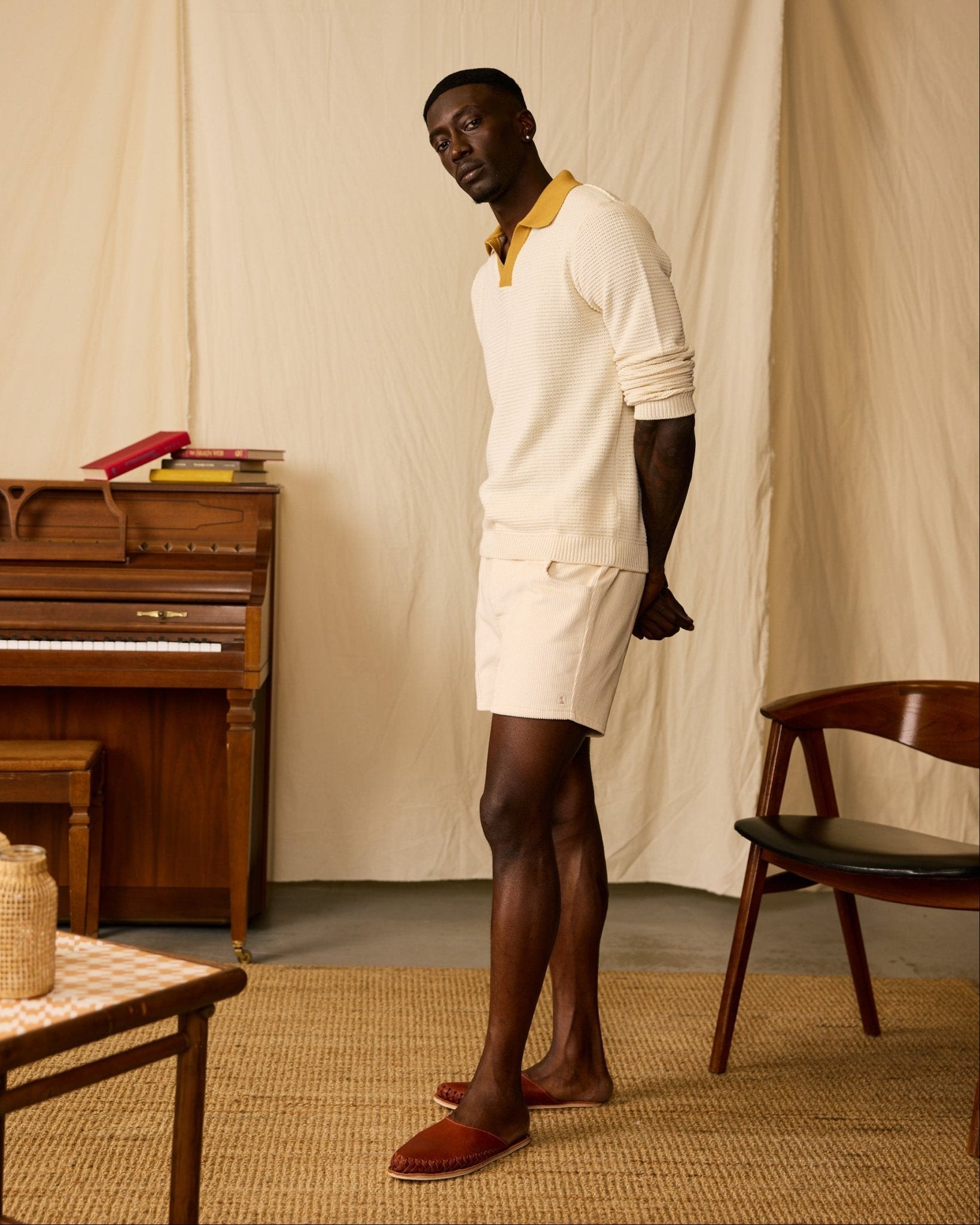 A man stands indoors on a woven rug, wearing a cream sweater and The Corsica Corduroy Shorts - Scallop by Dandy Del Mar with red slippers. A wooden piano, stacked books, a chair, and a table are in the background.