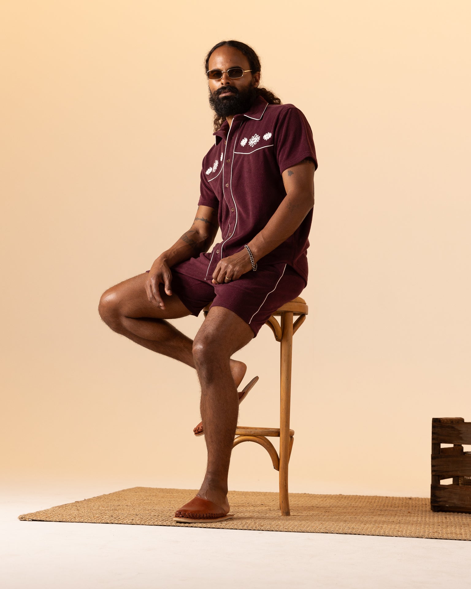 Donning The Gaucho Shirt in Bordeaux by Dandy Del Mar, a bearded person with sunglasses and a bun embodies an Equatorial Cowboy vibe. They sit confidently on a wooden stool against a beige backdrop, wearing soft terry cloth shorts.