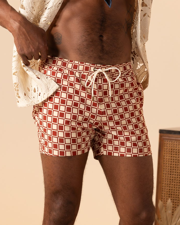 A person wears Dandy Del Mar’s The Ventura Volley Shorts - Rouge, featuring red and white checks with a white drawstring, paired with a cream openwork shirt—an effortless men's swimwear look against a neutral background.