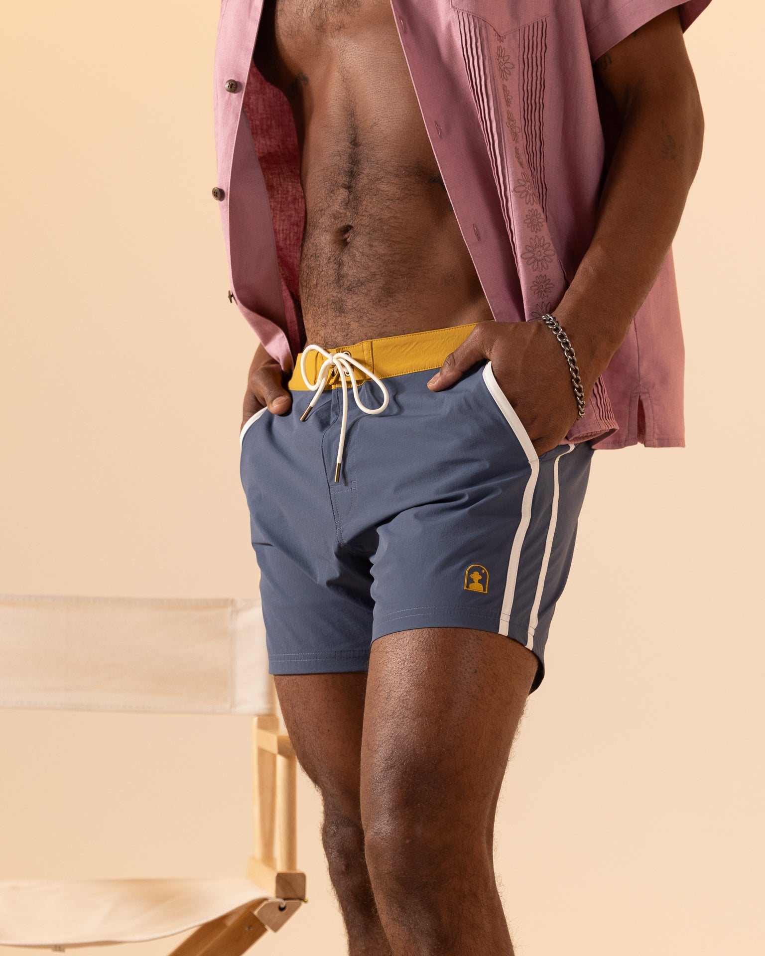 A person wearing a pink shirt and The Stirata Swim Short - Moontide by Dandy Del Mar stands near a beige chair.
