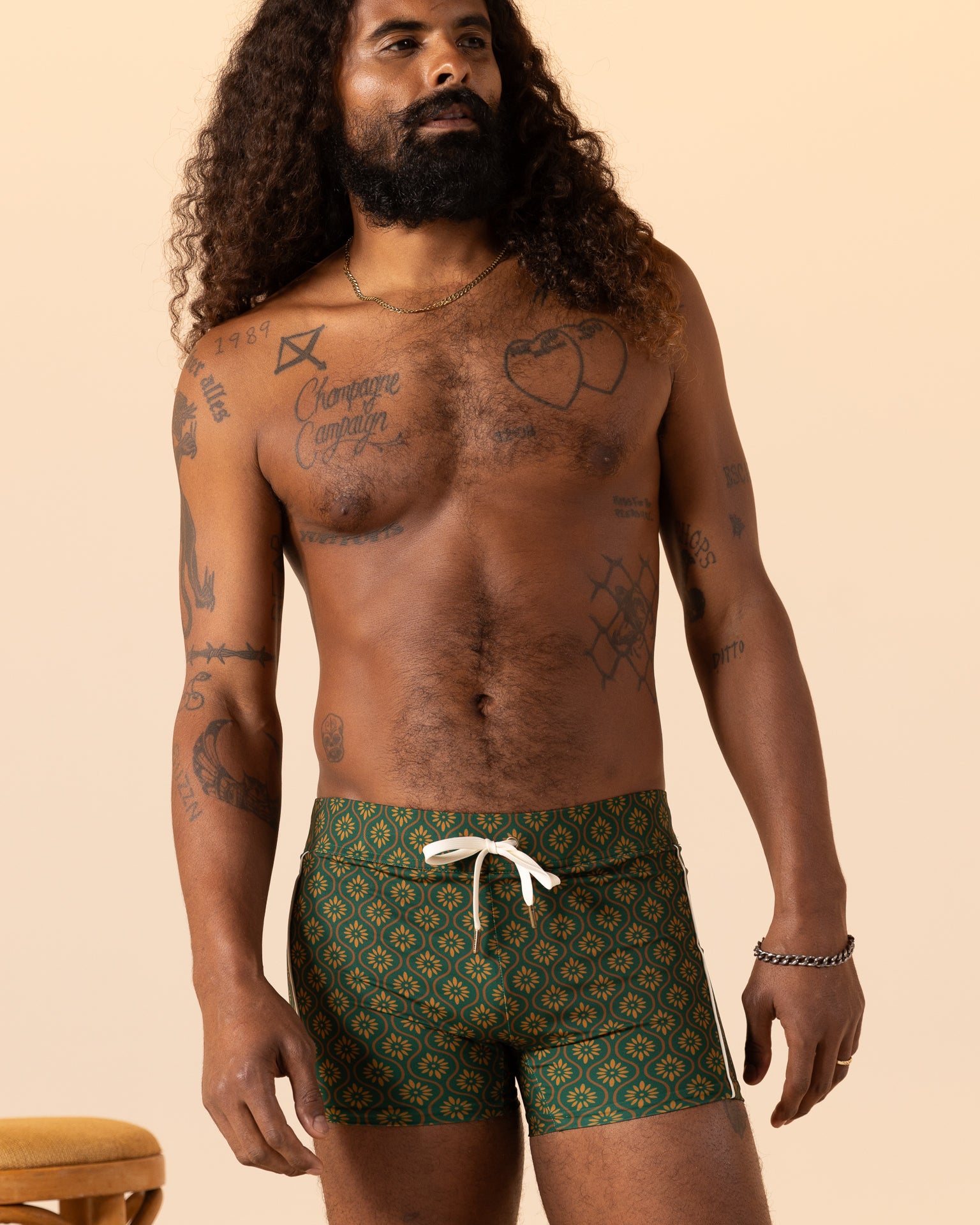 A man with long hair and tattoos models The Cassis Square Cut Swim Brief - Albero by Dandy Del Mar, displaying a European fit against a beige background.