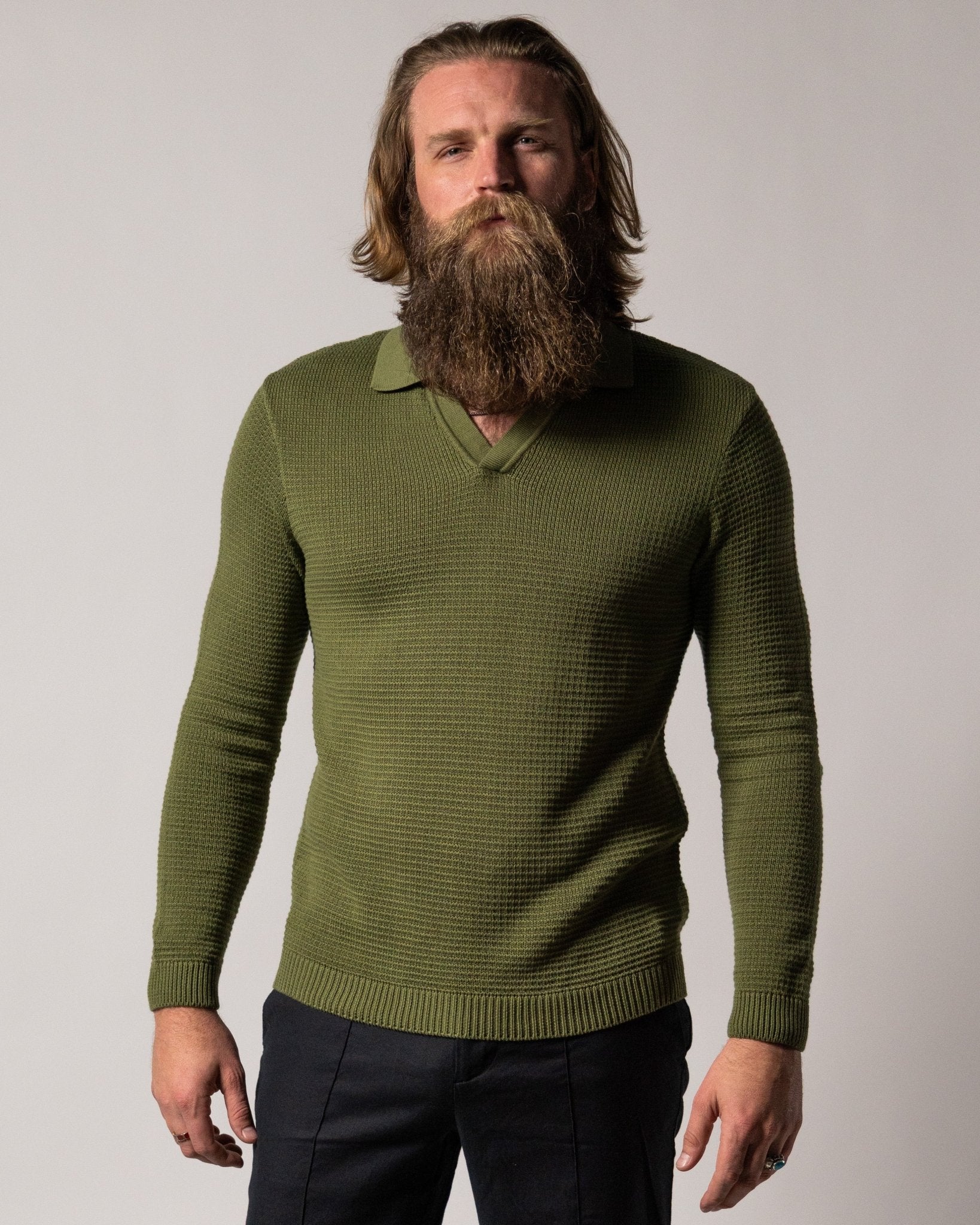 A man with long hair and a full beard wears the Dandy Del Mar Sebastian LS Polo Shirt in Arbequina, paired with dark pants, standing before a plain light background.