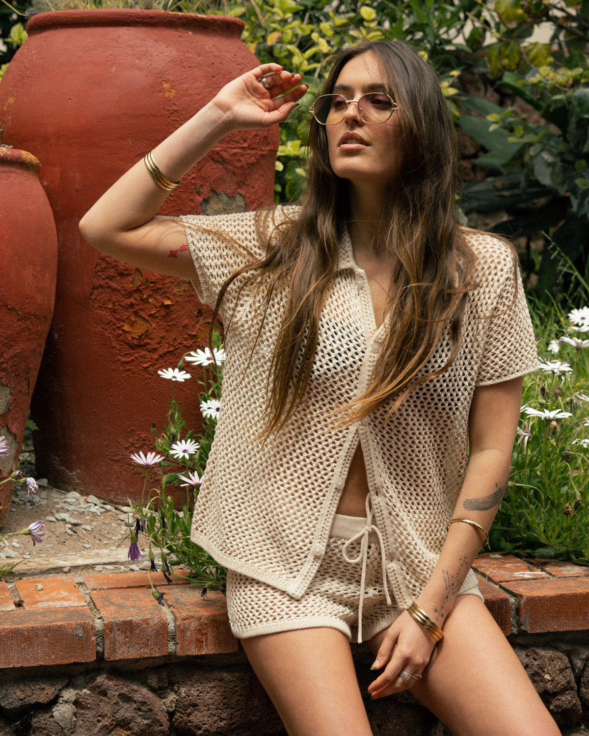 A woman in the Dandy Del Mar Malaga Crochet Shirt in Vintage Ivory relaxes on a brick ledge near terracotta pots and white flowers, sunglasses in hand, showcasing a summer-ready, effortless style.