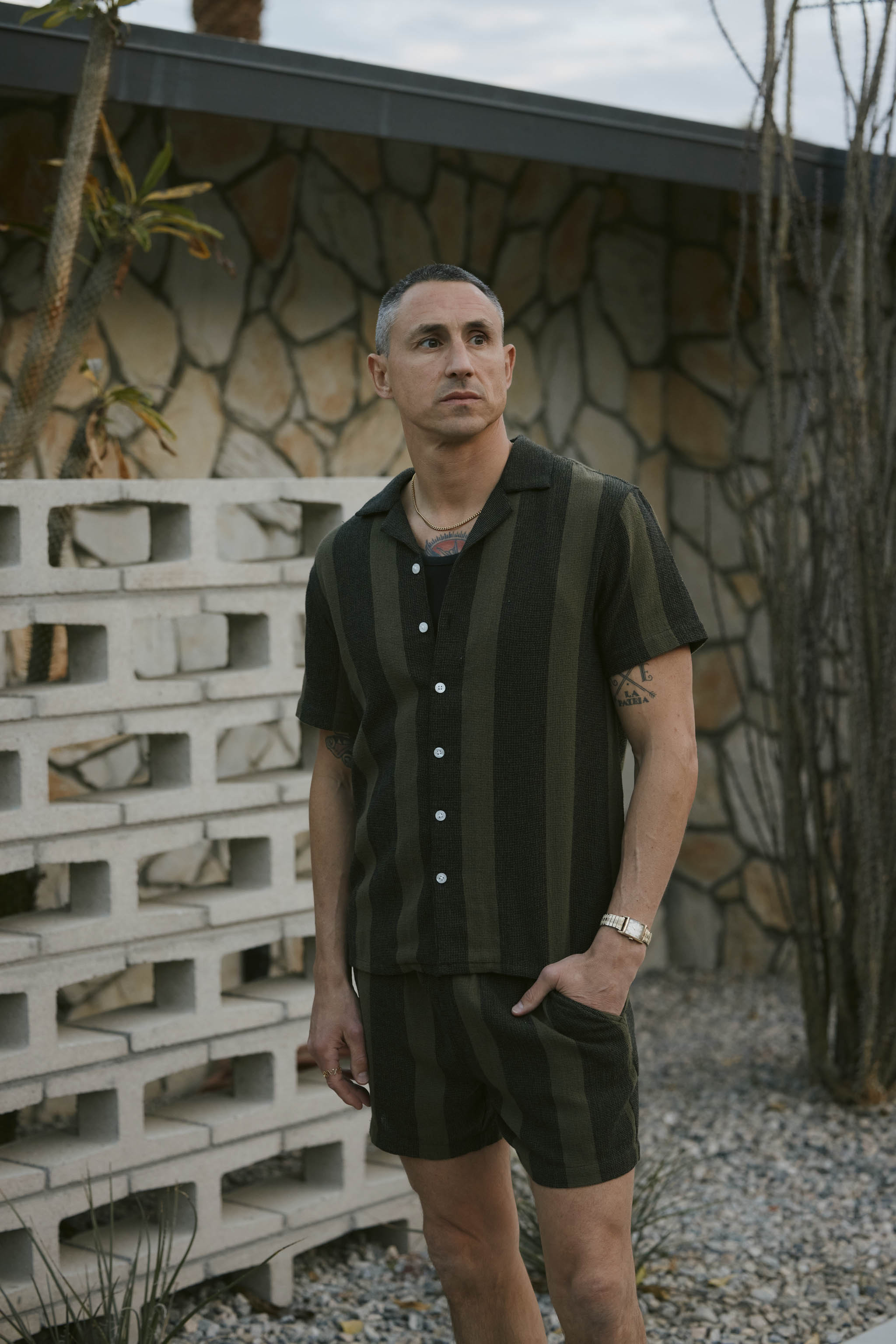 A person in a striped shirt and The Palma Short - Onyx by Dandy Del Mar stands outside near a stone wall with lush plants, evoking the aroma of exotic spices.