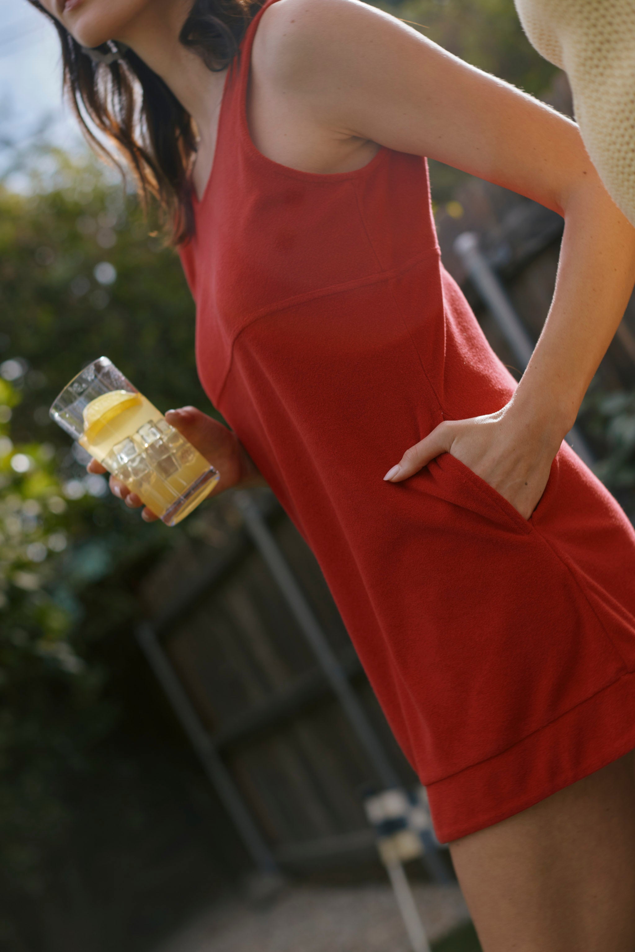A person wearing Dandy Del Mar's The Cyprus Dress - Pico, a red sleeveless design, holds a glass of lemonade with lemon slices in an outdoor setting.