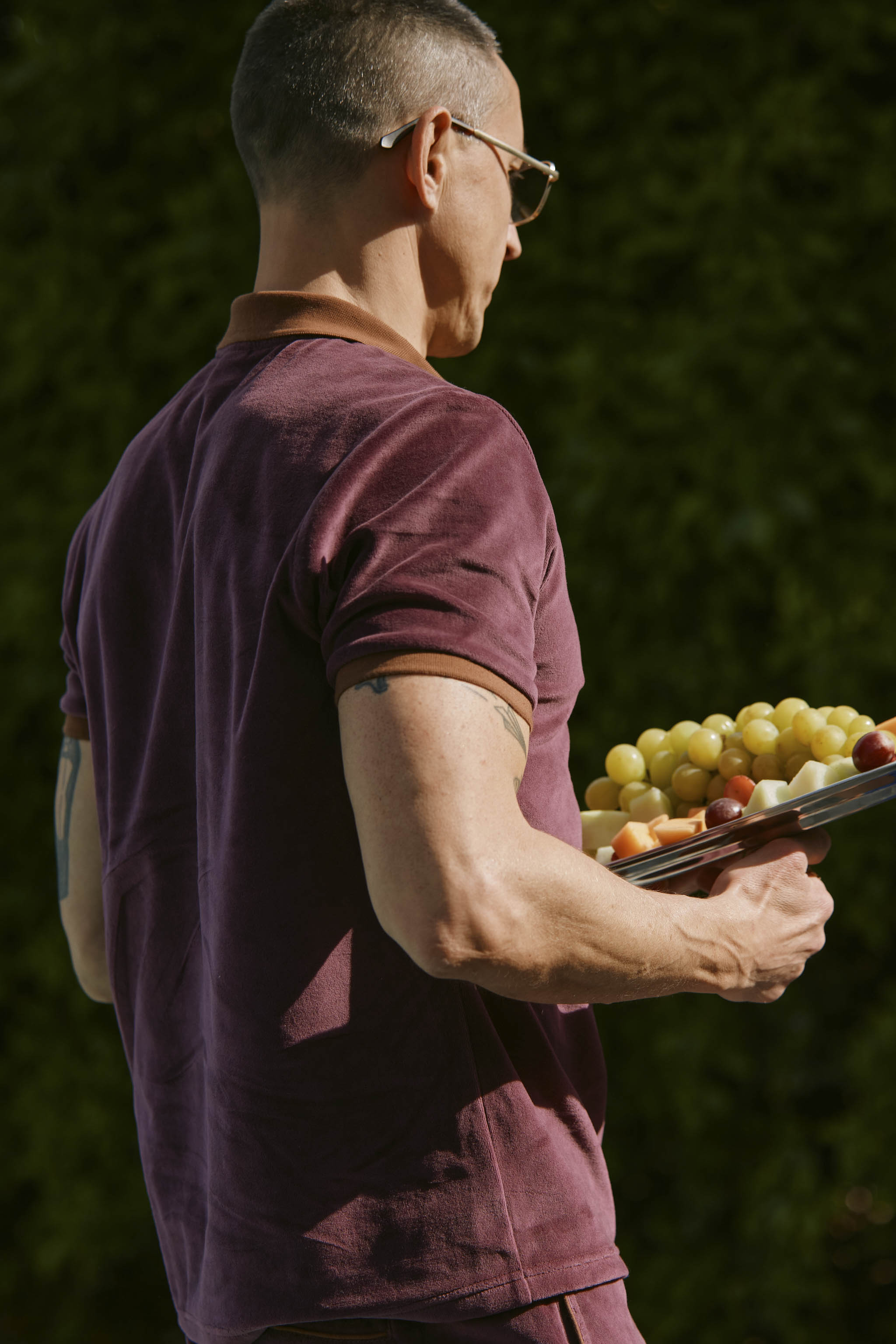 A person wearing The Rio Velour Shirt - Bordeaux by Dandy Del Mar holds a plate of green and red grapes.