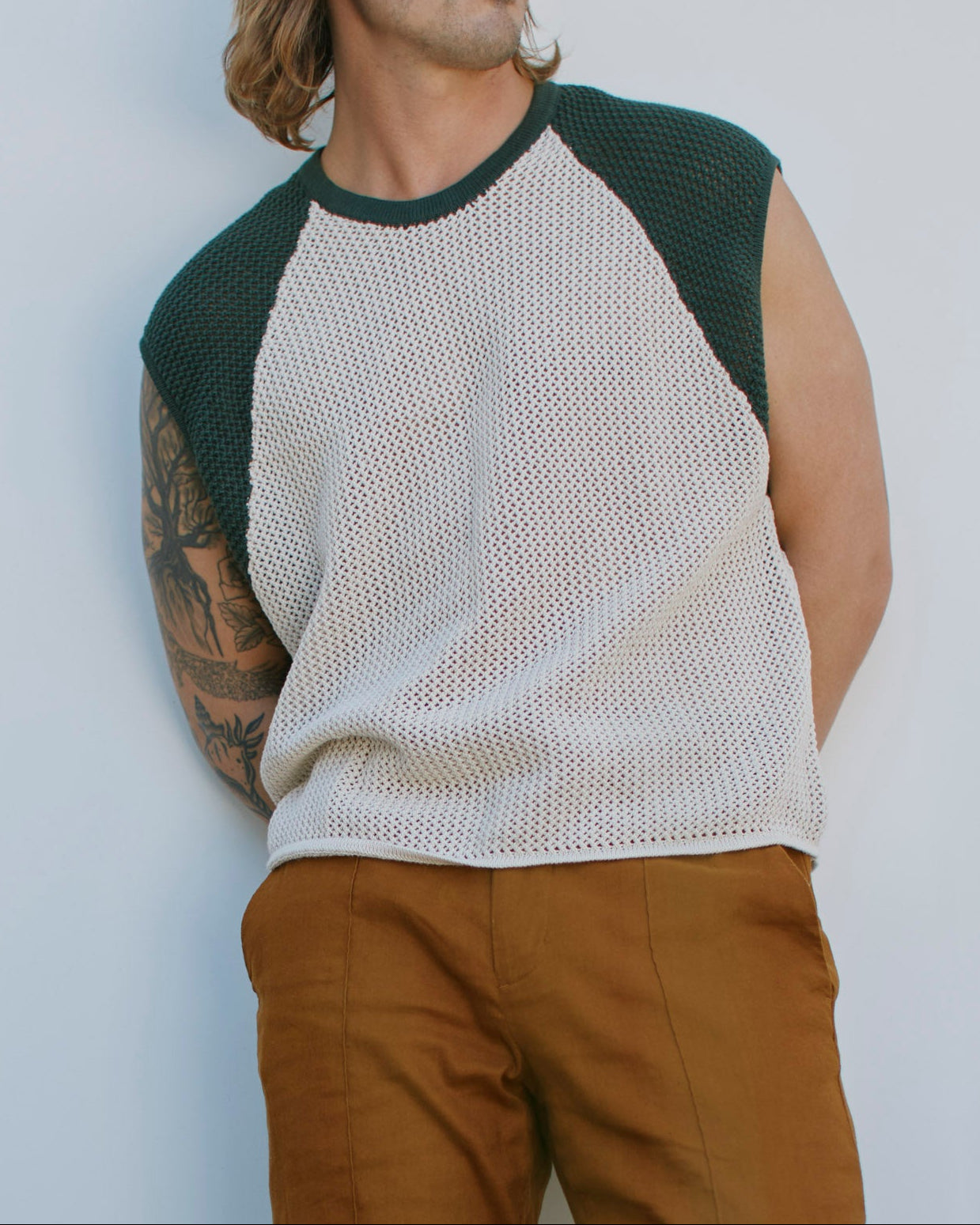 A man with shoulder-length hair and an arm tattoo wears The Porto Crochet Tank in Sabbia by Dandy Del Mar, styled with dark green sleeves and brown pants, posing against a plain light background.