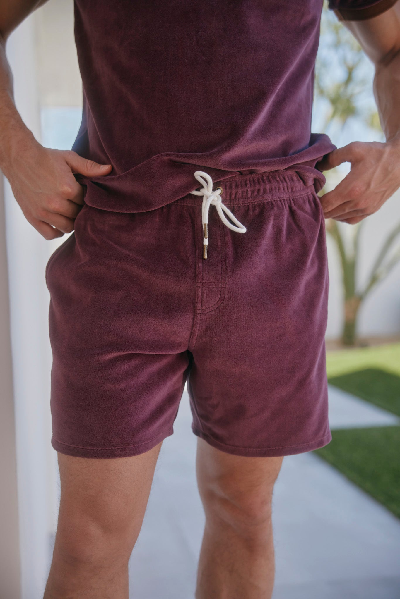 A person stands outdoors near a building wearing The Rio Velour Short - Bordeaux by Dandy Del Mar, featuring a white drawstring and sideseam piping, paired with a matching shirt.