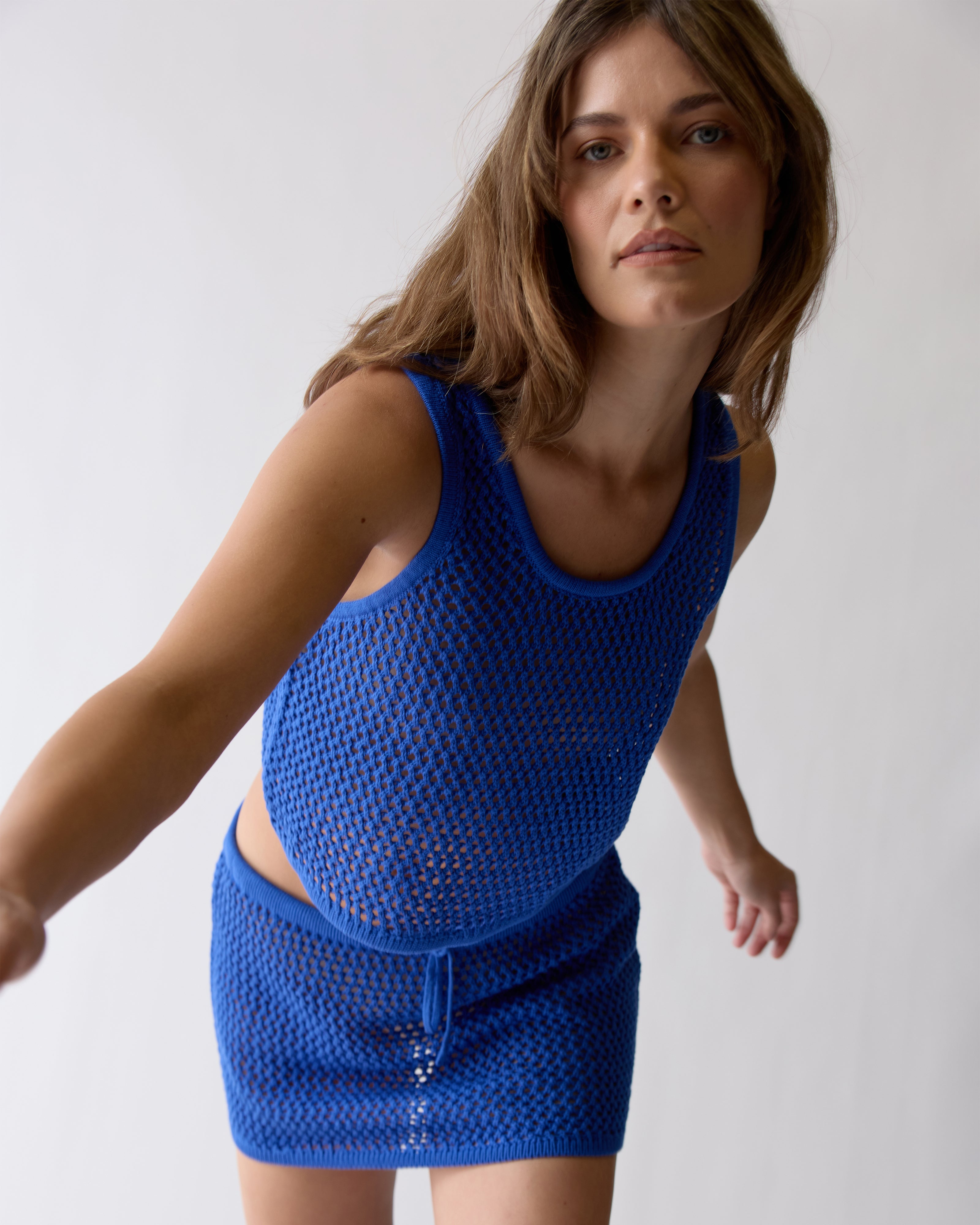 A woman in the Dandy Del Mar Malaga Crochet Tank - Solana, a 100% cotton cropped crochet tank, paired with a matching mini skirt, stands against a plain light background and leans slightly forward.