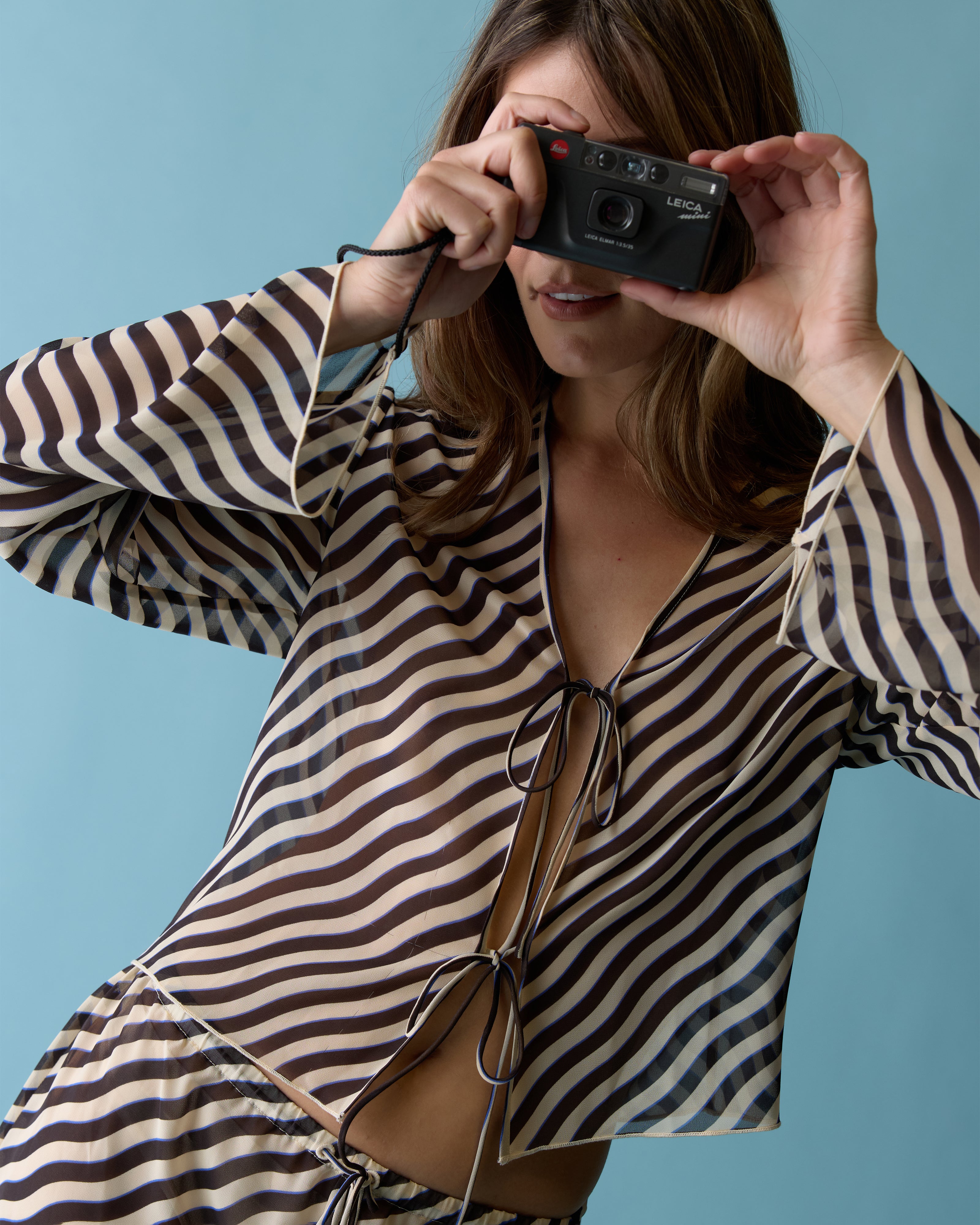 A woman in The Curacao Top - Tierra by Dandy Del Mar holds a camera up to her face while standing against a light blue background.