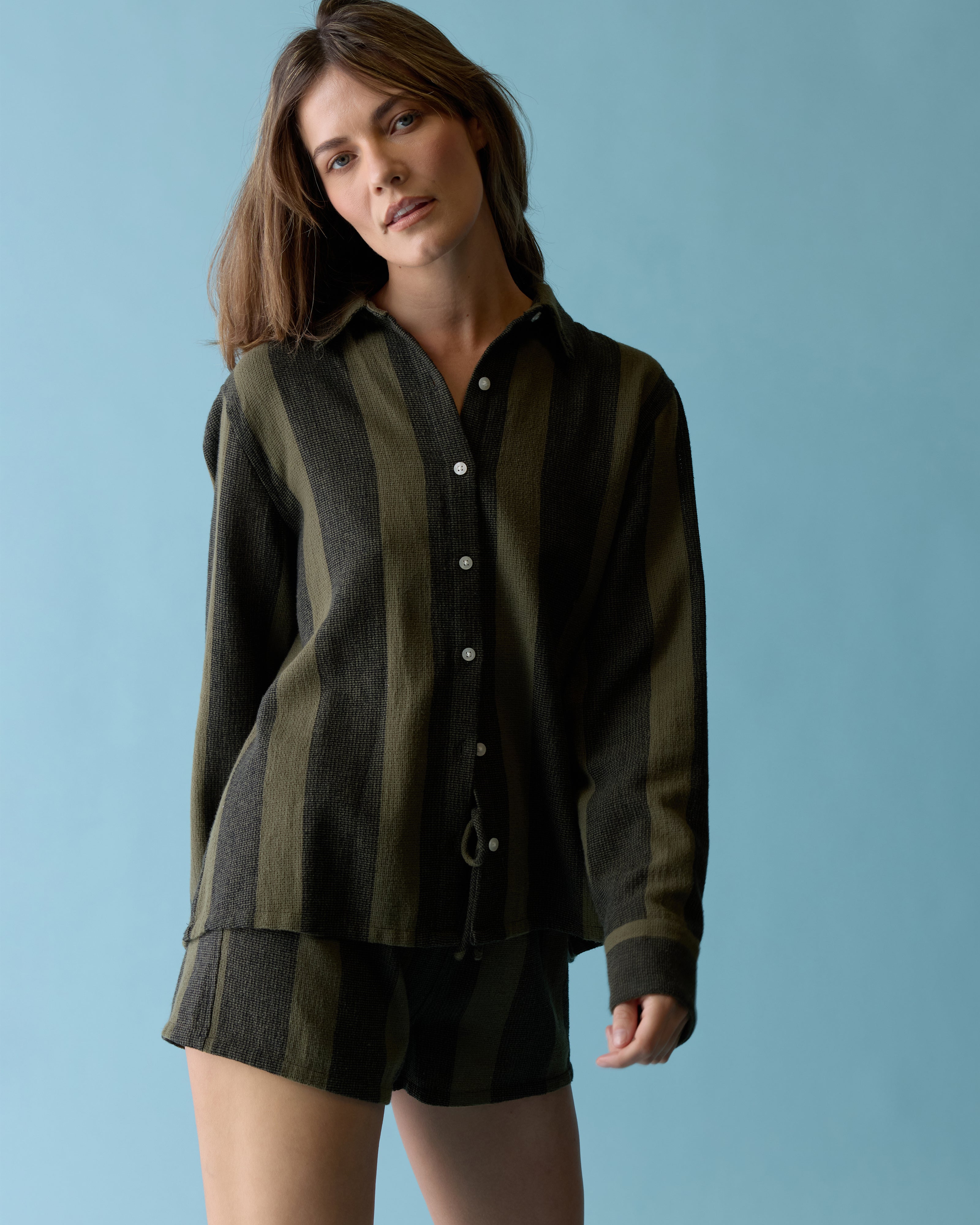 A person stands against a light blue background wearing The Sevilla Top - Onyx by Dandy Del Mar, a long-sleeve, button-up shirt with black and olive stripes.