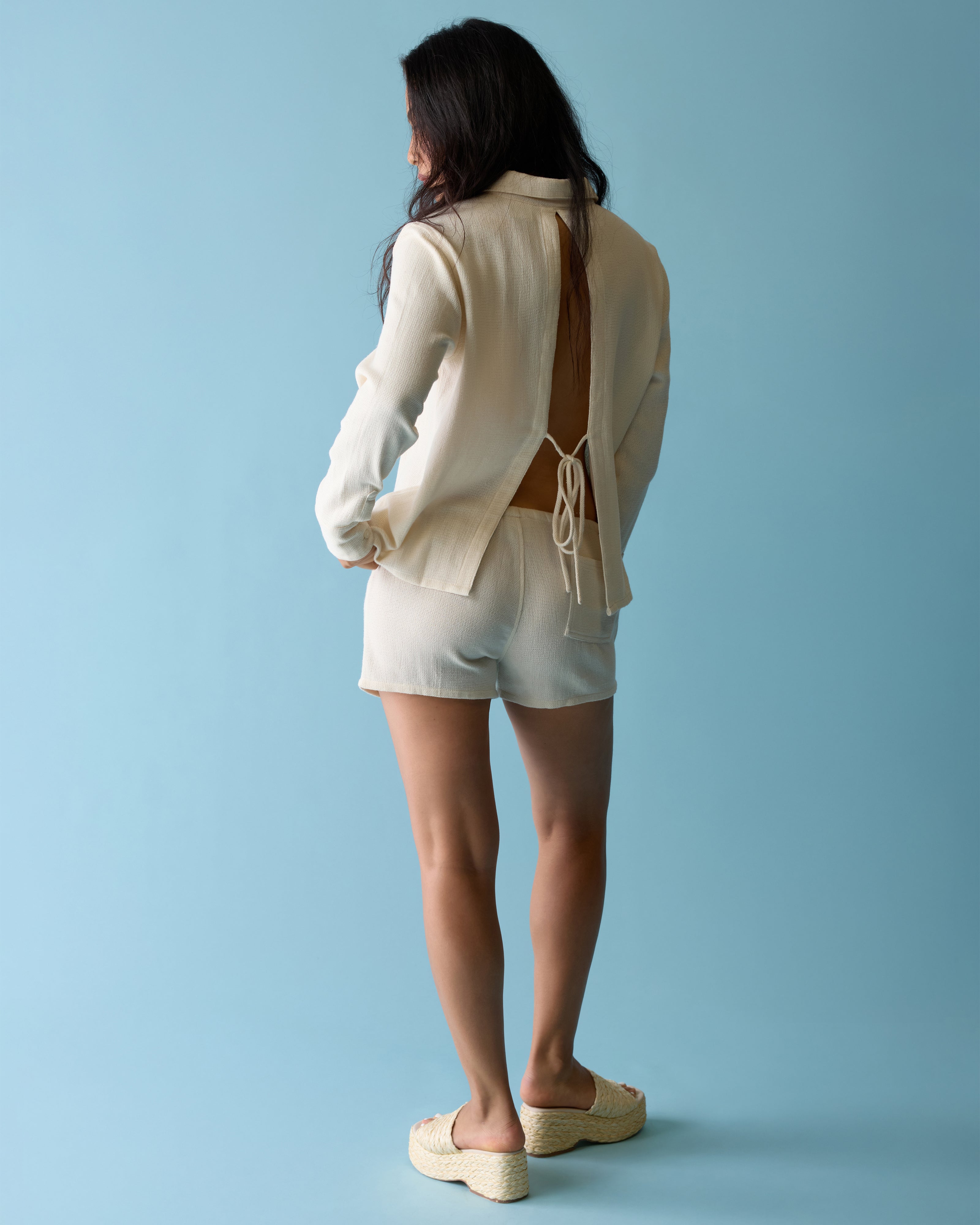 A person stands against a blue background wearing Dandy Del Mar’s Sevilla Top - Scallop, a cream-colored long-sleeve open-back top, paired with matching shorts and platform sandals.