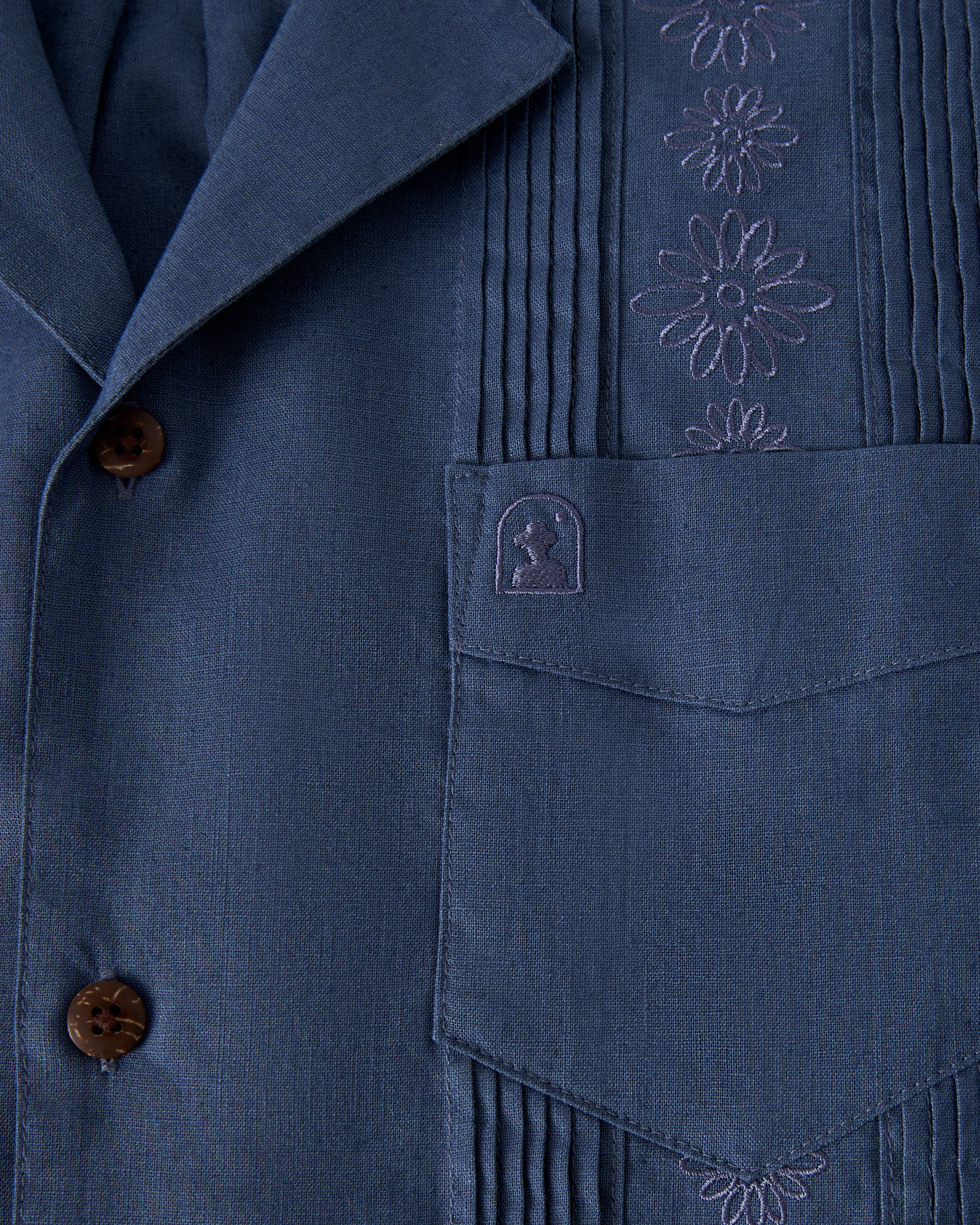 A close-up of The Brisa Linen Shirt - Moontide by Dandy Del Mar, featuring blue fabric with floral embroidery and vertical pleats. It includes brown buttons, a front pocket, and a small embroidered logo.