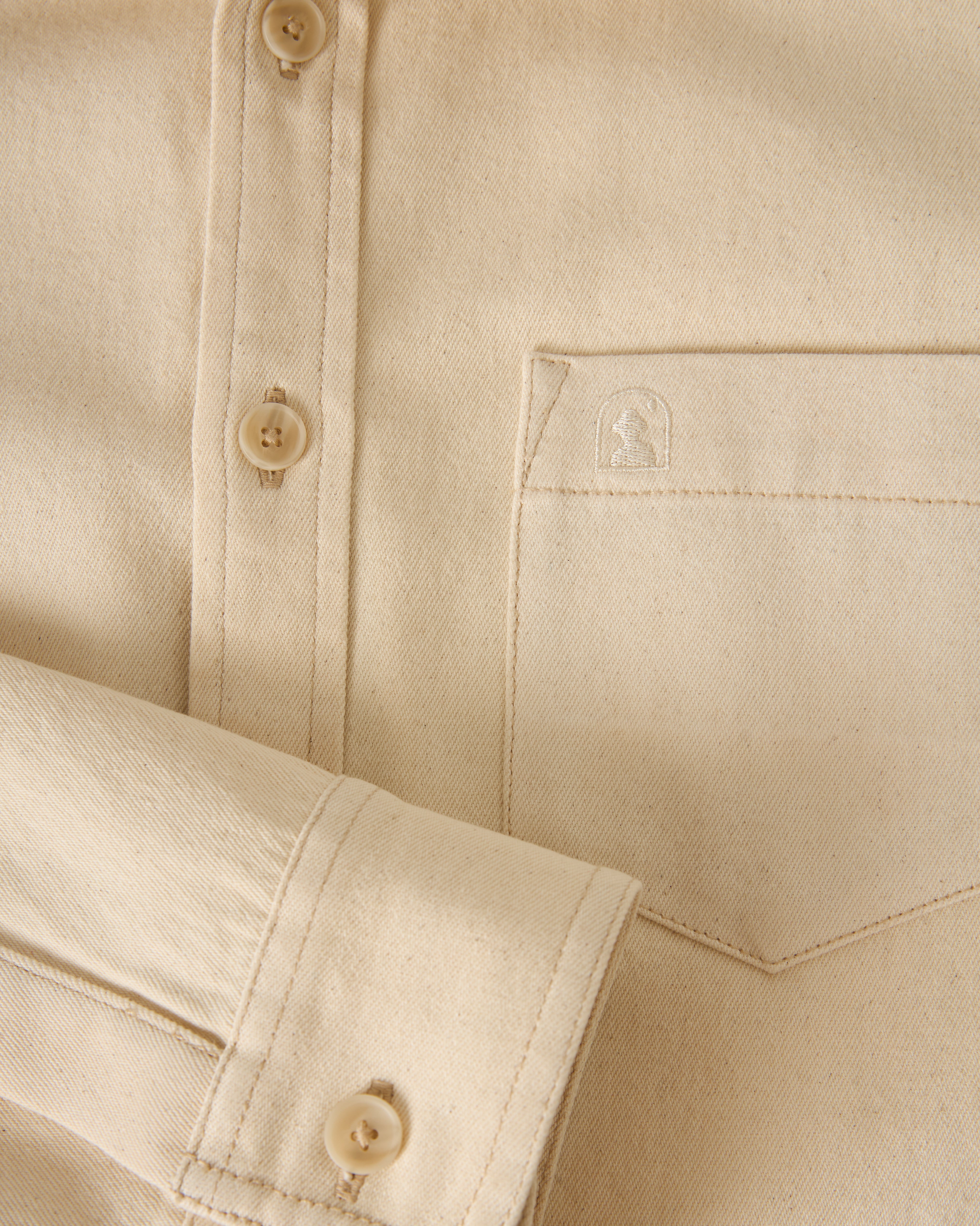 Close-up of The Cartagena Denim Shirt - Scallop by Dandy Del Mar, featuring a beige button-up design with a front pocket and a visible brand logo stitched on the pocket.