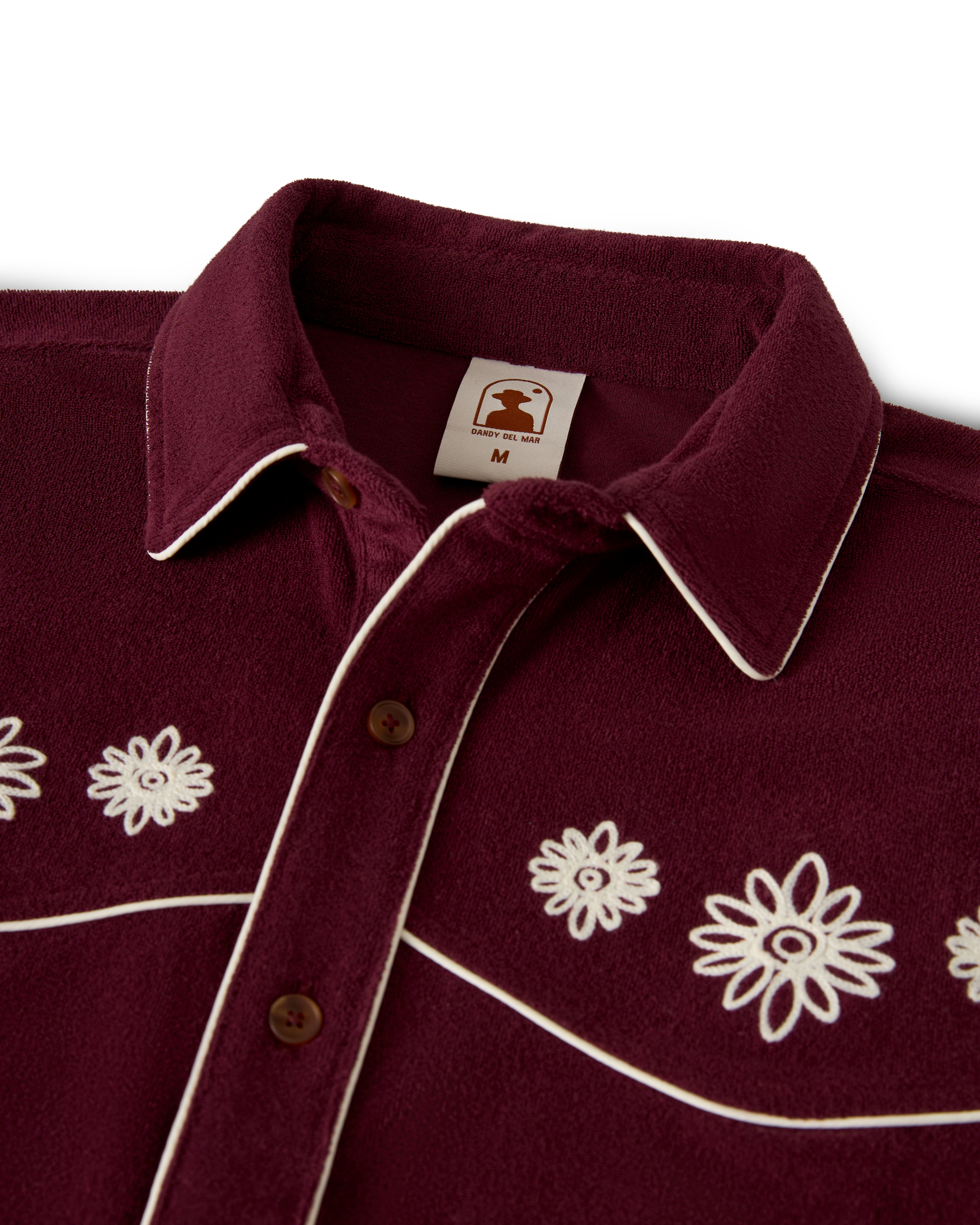 Close-up of The Gaucho Shirt - Bordeaux by Dandy Del Mar featuring floral embroidery and brown buttons. White piping outlines the collar and button area for an Equatorial Cowboy vibe. Inside, a label indicates size "M.
