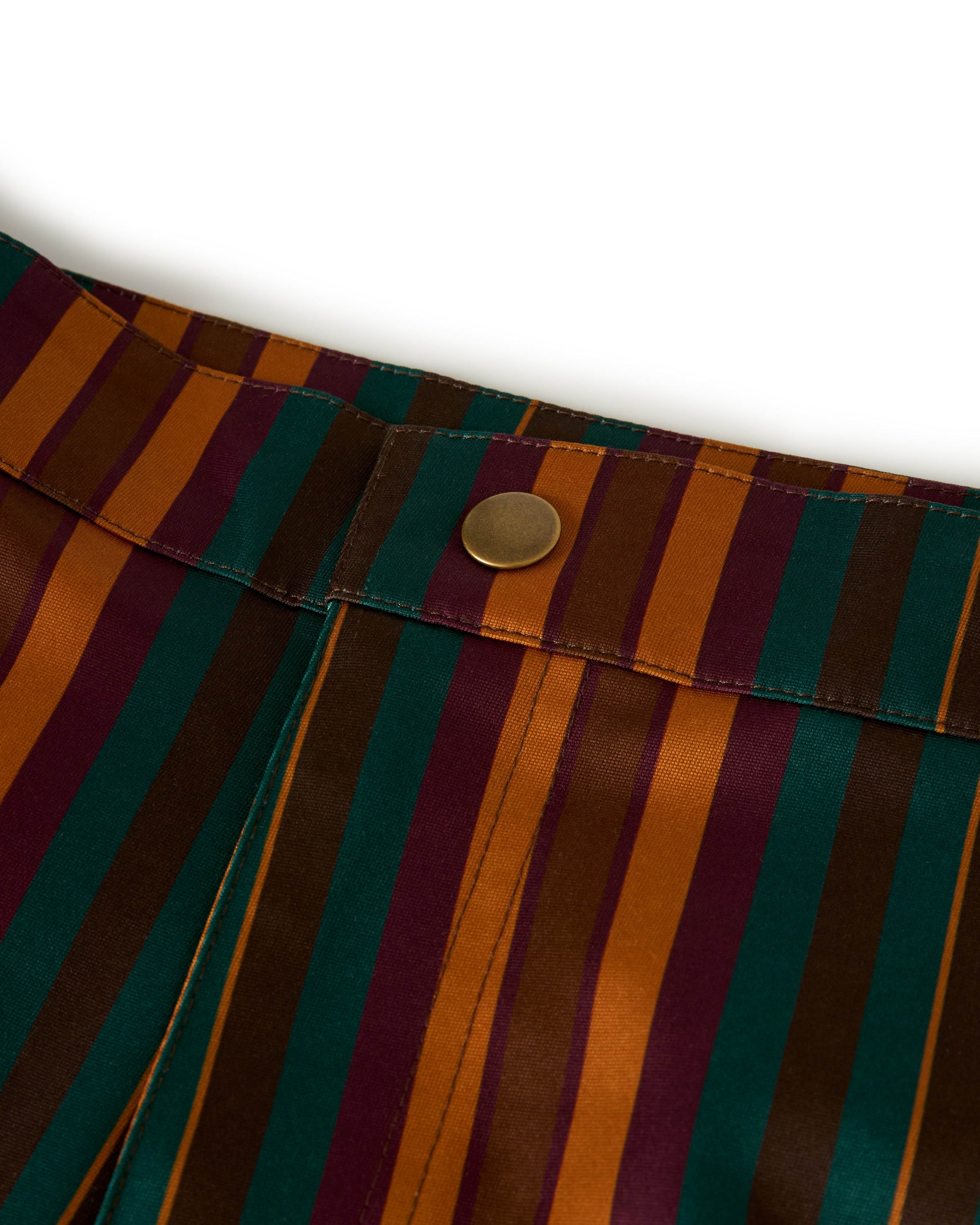 The Dandy Del Mar Mallorca Swim-Walk Short in Burnt Sienna features a close-up of striped fabric in orange, green, purple, and brown tones with a gold button on the waistband.