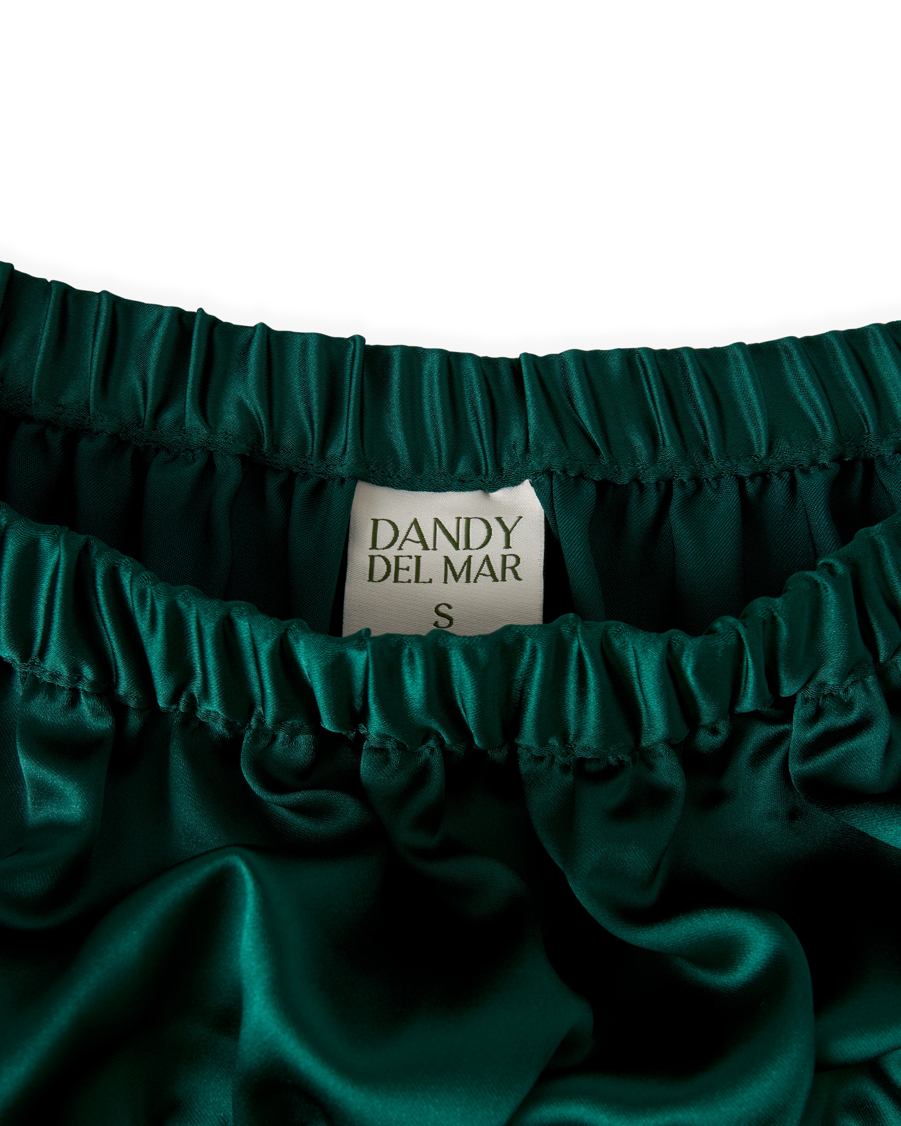 Close-up of a dark green satin garment with an elastic waistband, displaying a label reading "The Monaco Maxi Skirt - Albero by Dandy Del Mar, Size S".