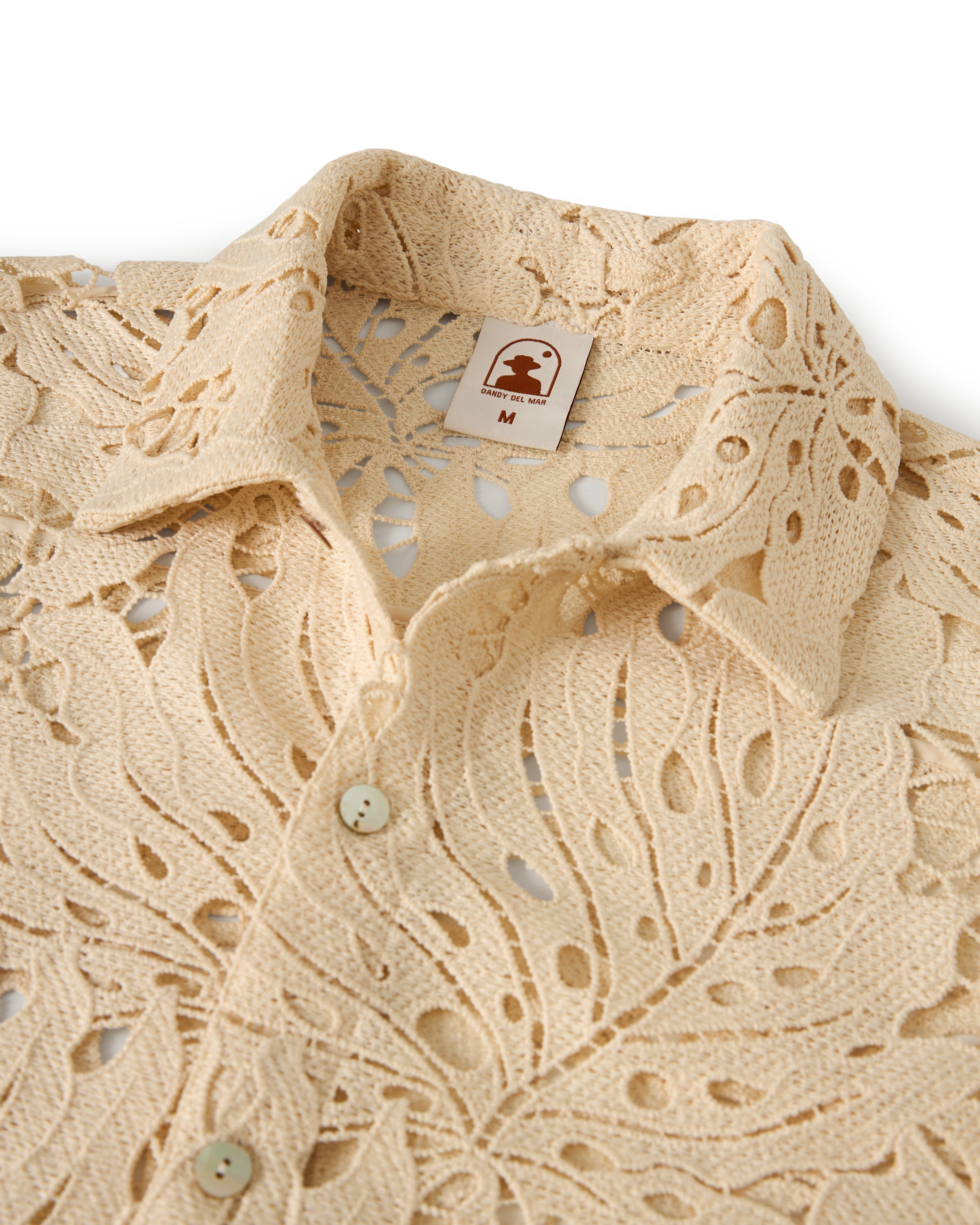 The Monstera Shirt - Scallop by Dandy Del Mar is a beige crochet lace button-up shirt with intricate floral patterns and a brand tag at the collar indicating size "M".