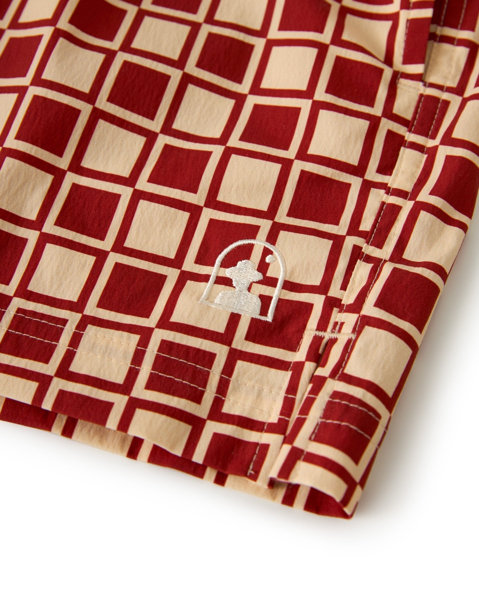 Close-up of The Ventura Volley Shorts - Rouge by Dandy Del Mar, showcasing red and beige checkered fabric, elastic waist, white embroidered abstract logo near the hem, and detailed stitching.