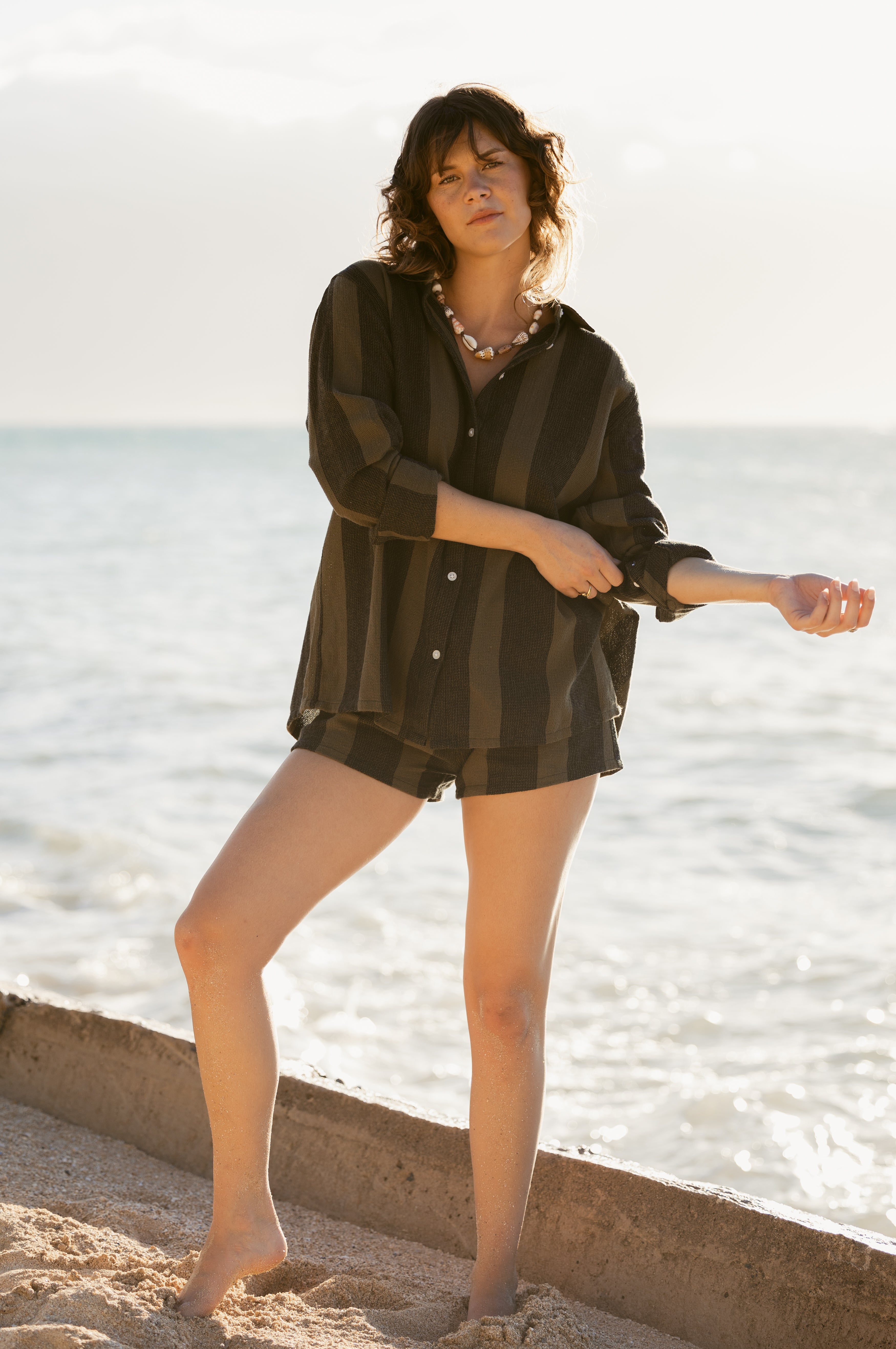 A woman stands barefoot on the sand in a loose, striped shirt and Dandy Del Mar's The Sevilla Short - Onyx, as the sea and sky extend behind her.