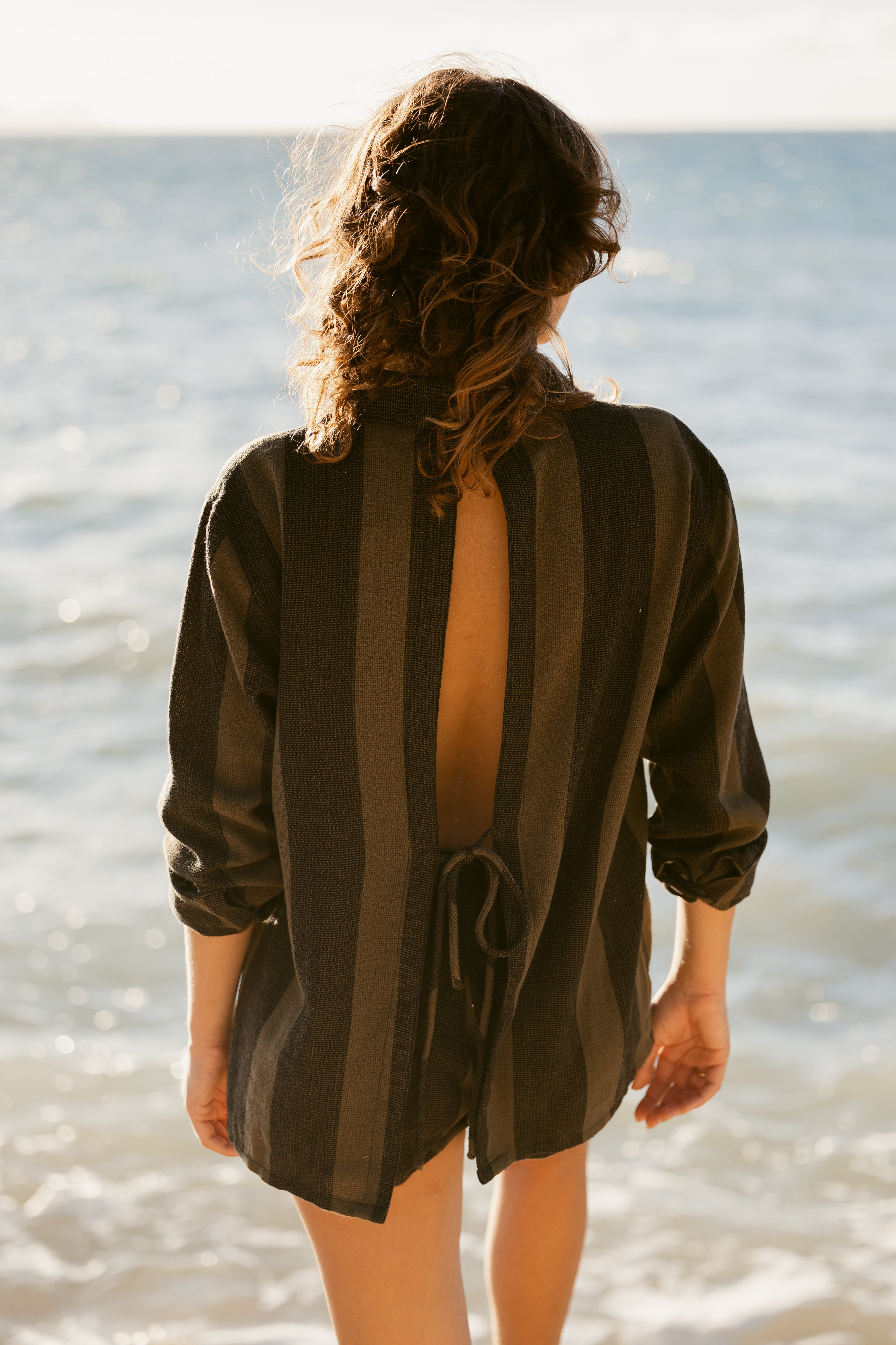 A person with curly hair stands on the beach facing the ocean, wearing Dandy Del Mar's Sevilla Top – Onyx, a resort-ready long-sleeve striped shirt featuring an open back and lower-back tie.