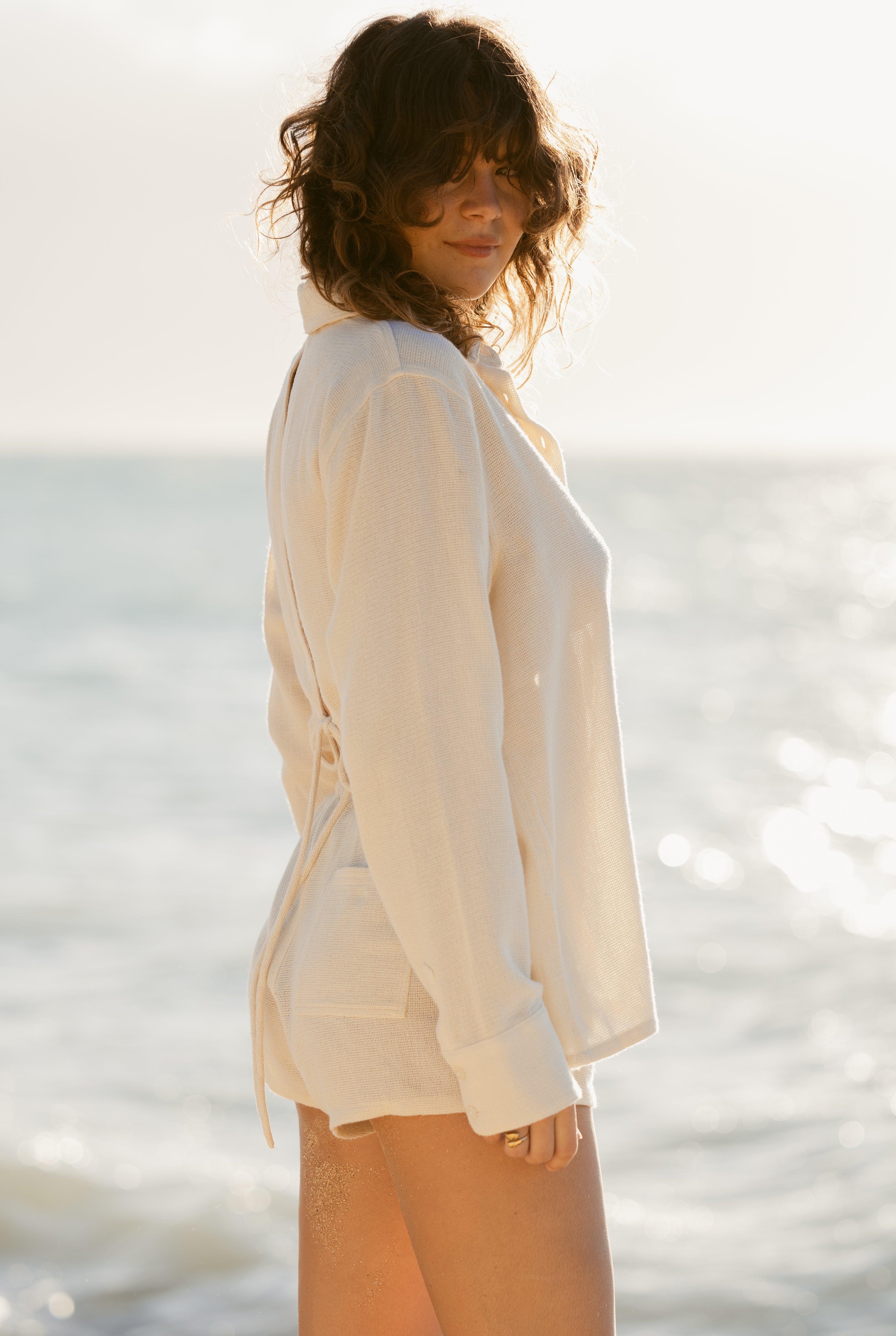 A woman with wavy hair, in a long-sleeved white shirt and Dandy Del Mar’s The Sevilla Short - Scallop with an elastic waistband, stands sideways on the beach under bright sunlight with the ocean behind her.
