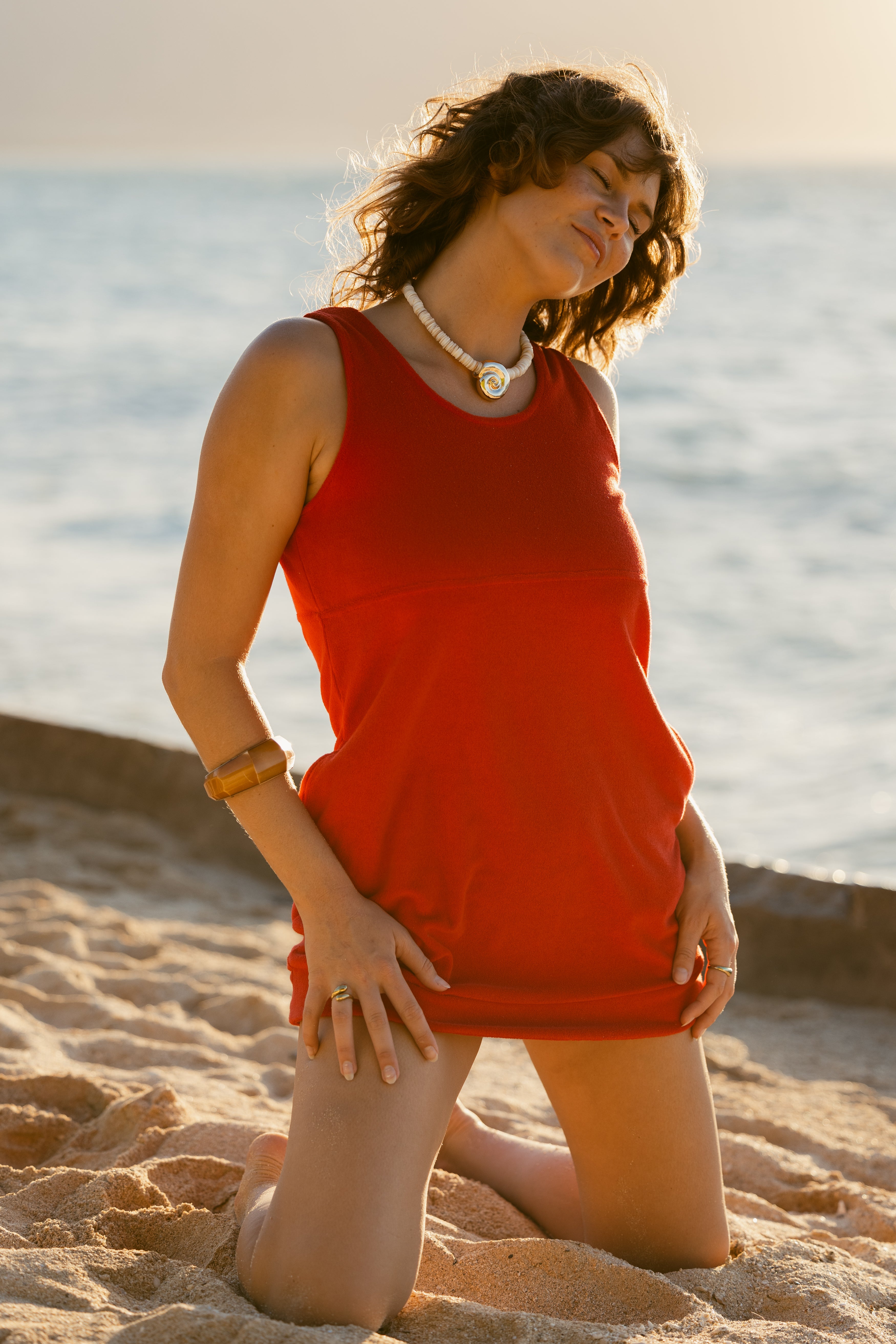 A person in The Cyprus Dress - Pico by Dandy Del Mar kneels on a sandy beach near the ocean, sunlight illuminating the scene.