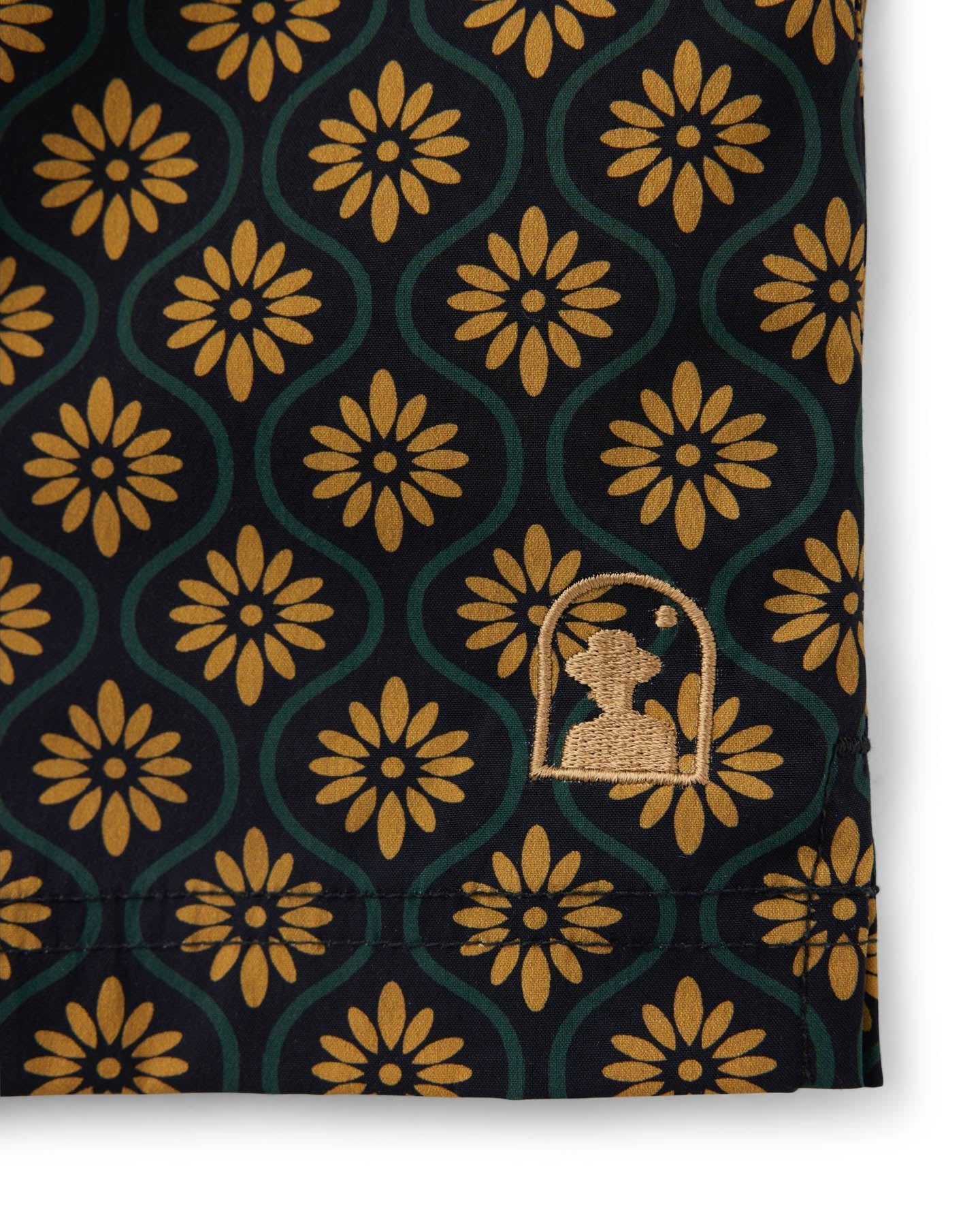 Close-up of The Ventura Volley Shorts - Oro Gardenia Print by Dandy Del Mar, featuring men’s swimwear fabric with a repeating gold floral pattern on a dark base, elastic waist, and a small stitched silhouette logo near the hem.