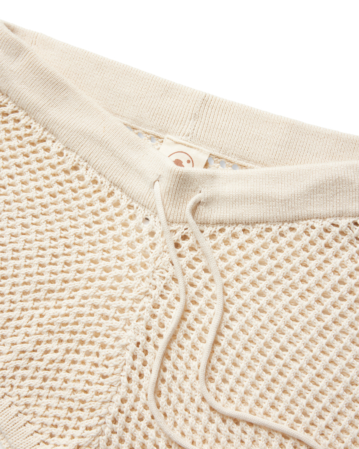 Close-up of The Malaga Crochet Shorts in Vintage Ivory by Dandy Del Mar, featuring a ribbed waistband with adjustable drawstring and a tag adorned with a simple smiley face design.