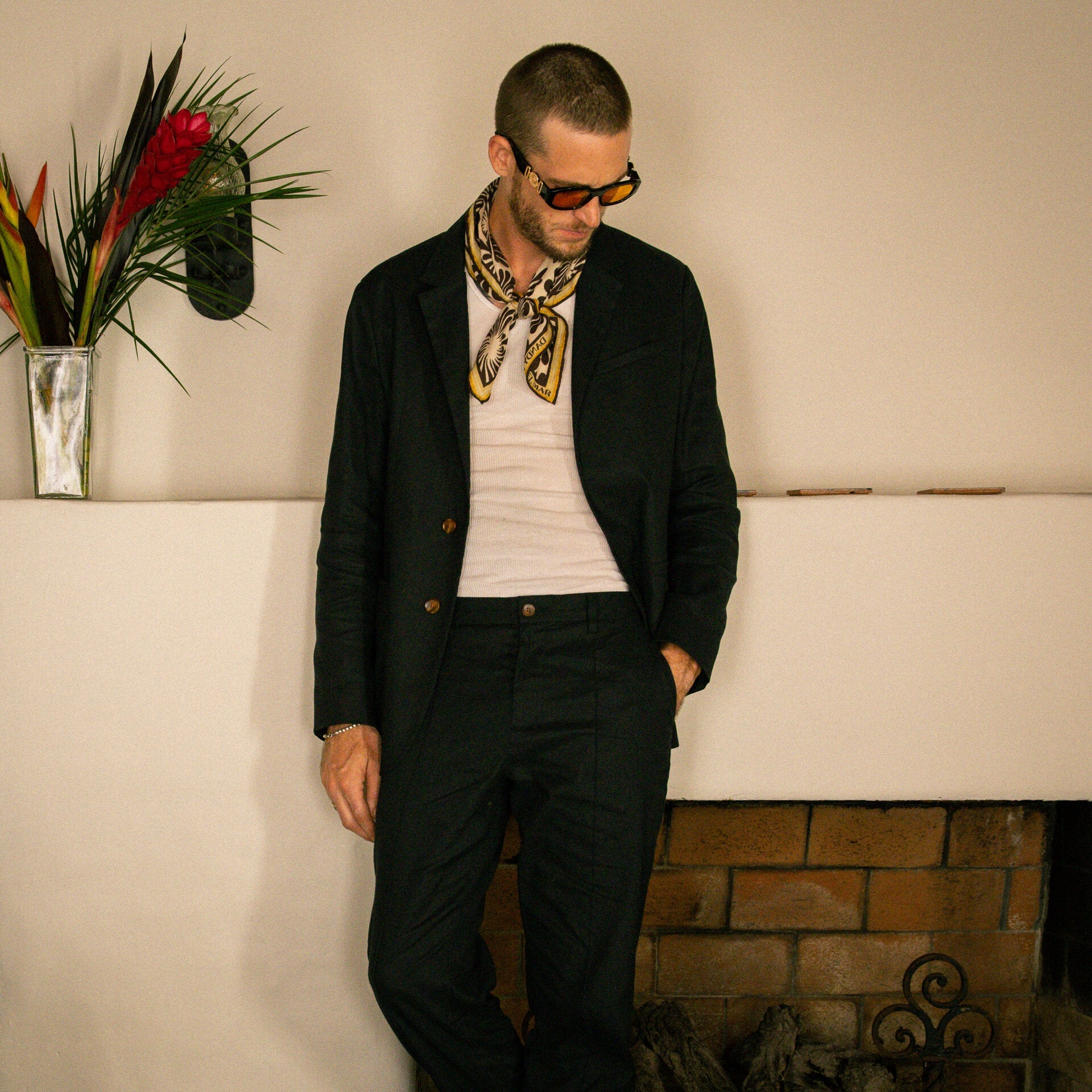 A man wearing sunglasses, a black suit, a white shirt, and a patterned scarf stands in front of a white mantel with a tropical flower arrangement.
