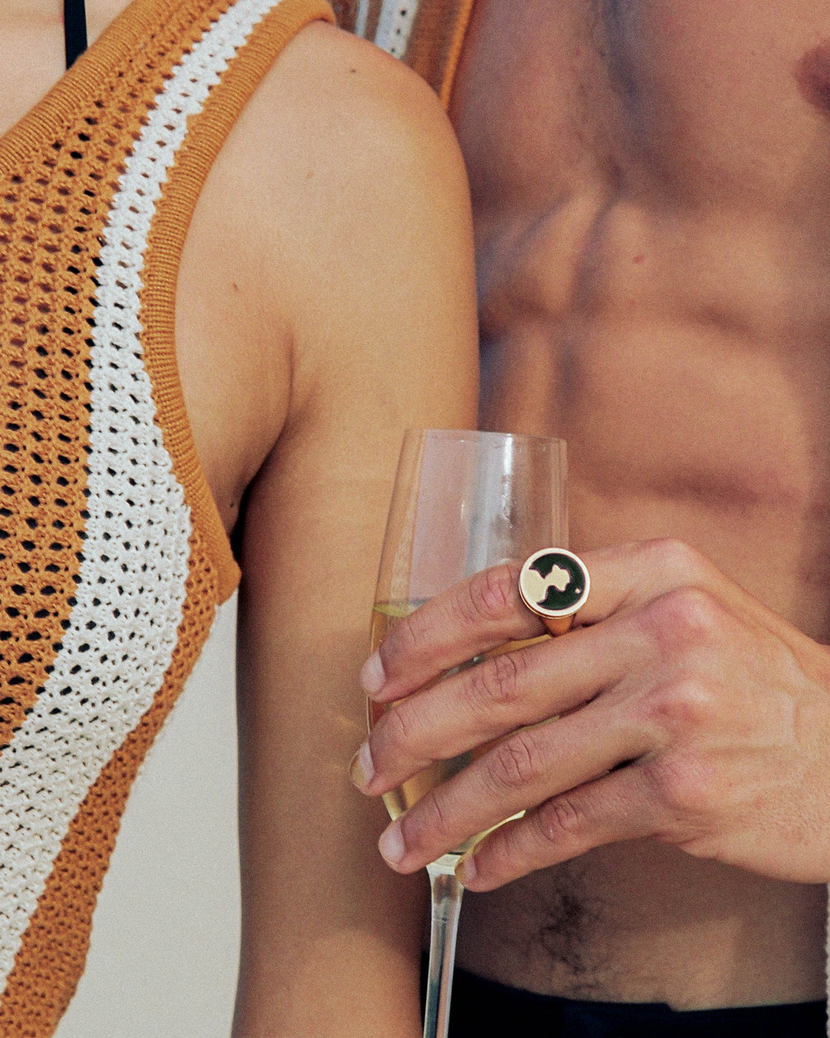 Two people standing close together, one wearing a crocheted top, the other shirtless, holding a champagne glass with a large round ring on their finger.