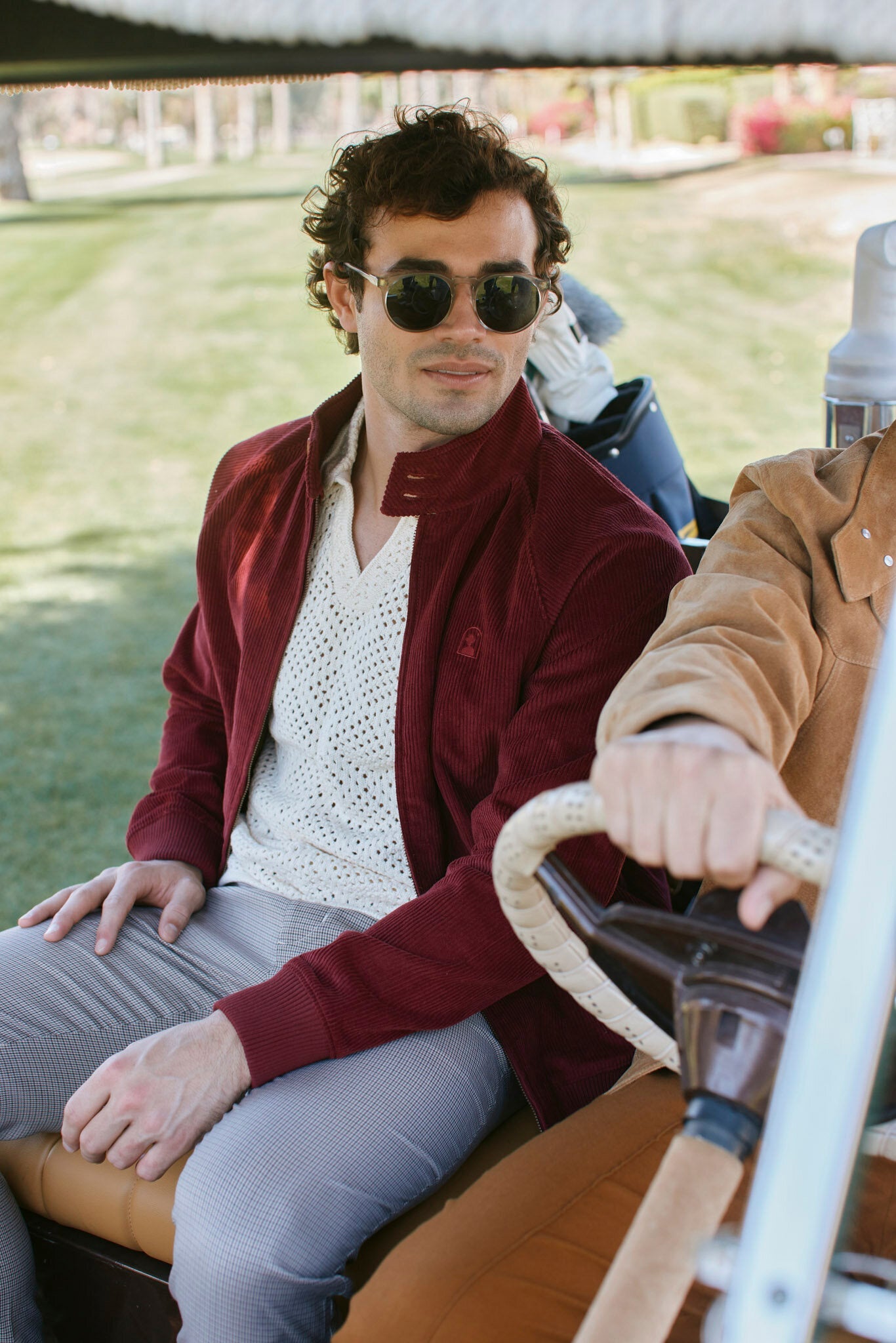 A man wearing sunglasses, a red jacket, and light pants sits beside the driver in a golf cart on a grassy area.