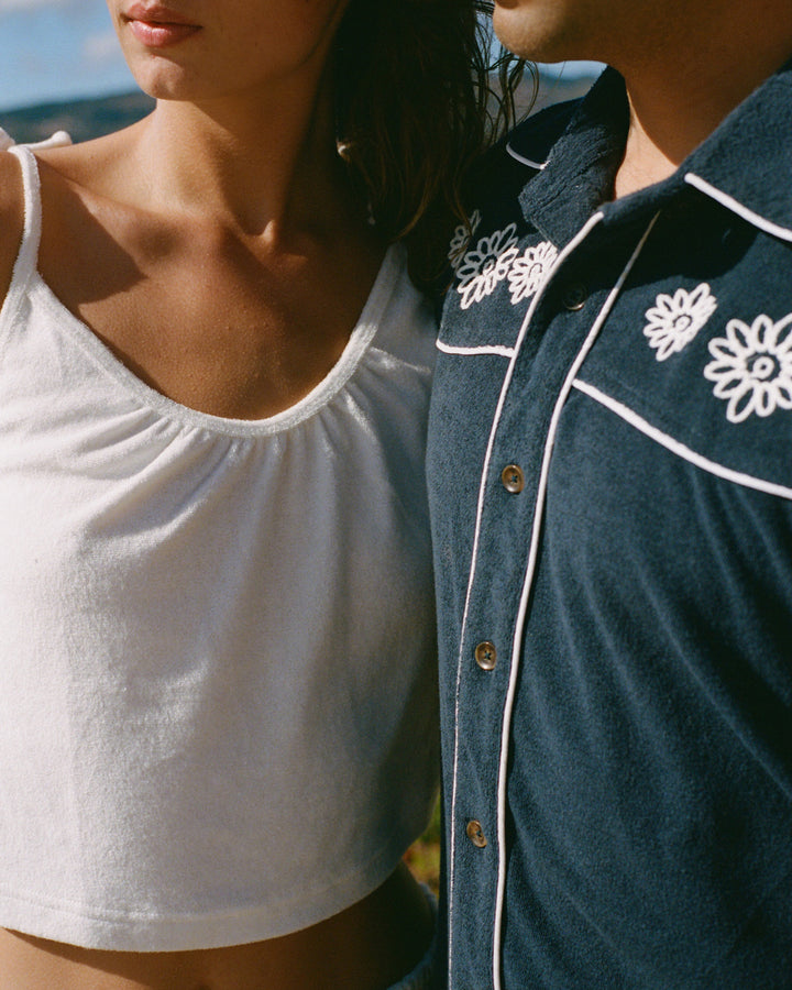 Close-up of a couple standing together. The woman wears a Vintage Ivory sleeveless La Jolla top with adjustable straps from Dandy Del Mar, while the man dons a navy shirt adorned with floral embroidery.