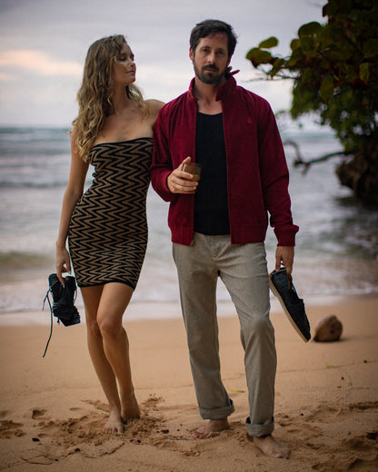 A woman in a Gardenia jacquard silk dress and a man in The Corsica Corduroy Jacket - Sangria by Dandy Del Mar stand barefoot on a beach, holding shoes. The ocean and greenery are in the background.