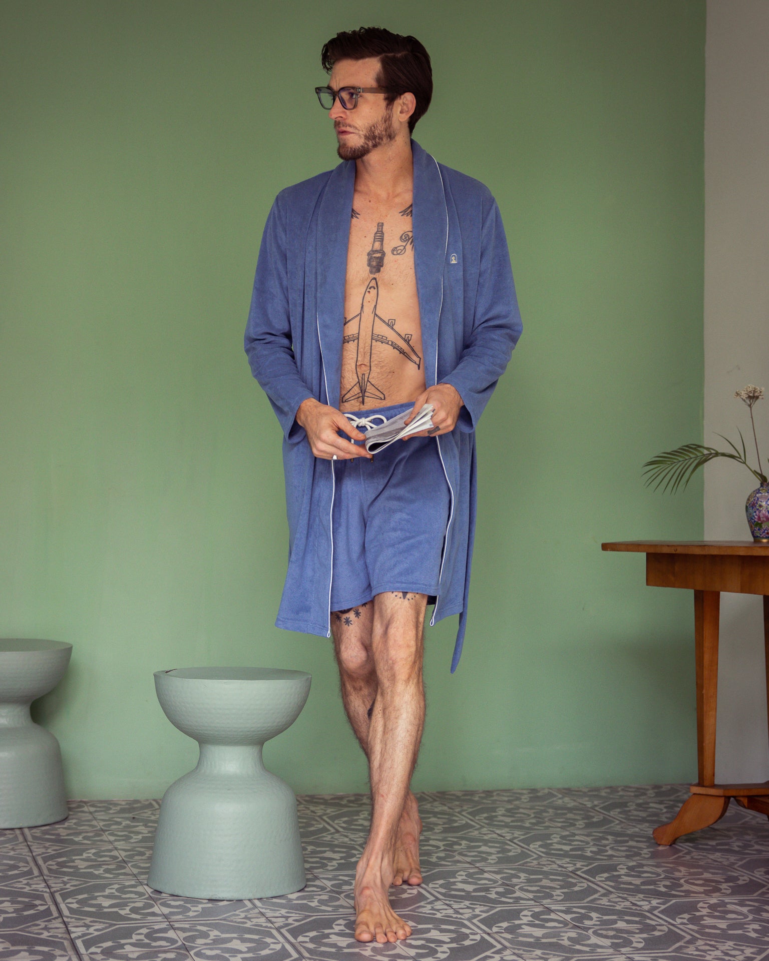 Clad in the ultra-soft blue Tropez Robe by Dandy Del Mar, the man stands on a patterned tile floor. He holds a book and glasses, with a vase-topped table and two stools behind him against the green wall.