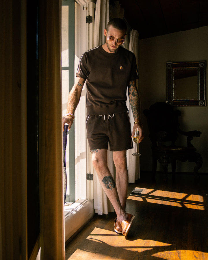 A man with tattoos standing in front of a window wearing the Marseille Pullover by Dandy Del Mar - Carajillo.