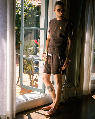 A man stands indoors by an open door, holding sunglasses and a tennis racket, wearing Dandy Del Mar’s The Marseille French Terry Shorts in Carajillo as sunlight streams in.