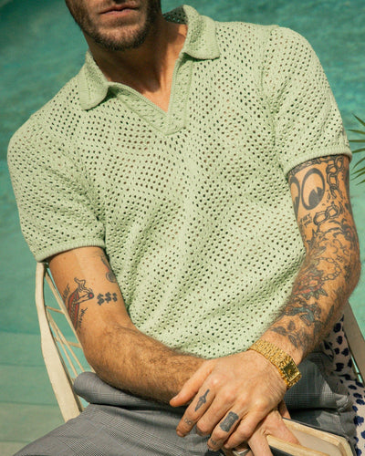 A tattooed man sits by a pool wearing the Antibes Crochet Polo Shirt in Pistachio by Dandy Del Mar, plaid pants, and a gold wristwatch.
