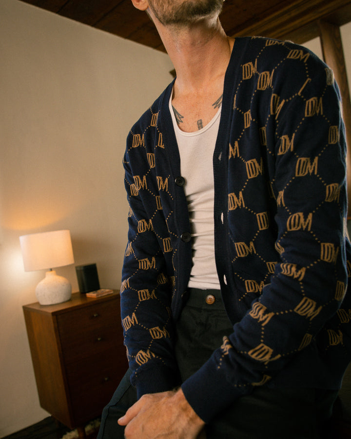 A man sitting on a bed wearing the Dandy Del Mar Castello Cardigan - Abyss.