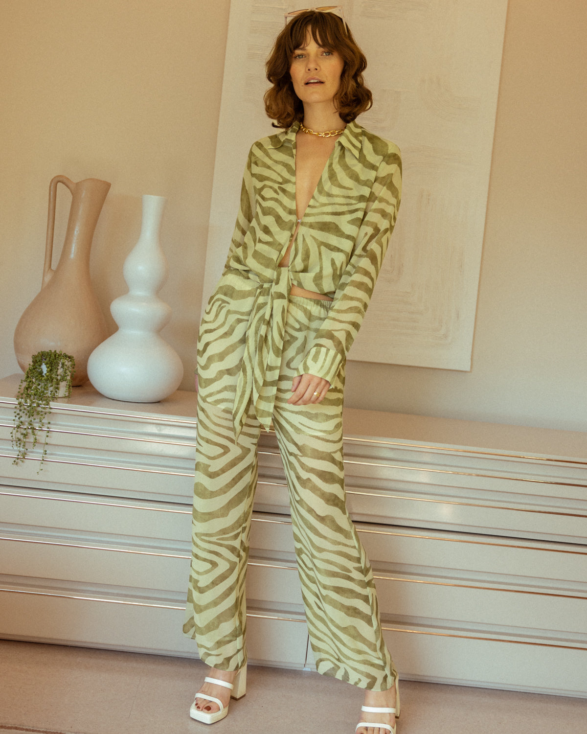 A person stands in a room wearing a vibrant green zebra-patterned outfit paired with white platform sandals, featuring the tie front design of The Cayman Top - Arbequina by Dandy Del Mar, in front of a dresser adorned with decorative vases.